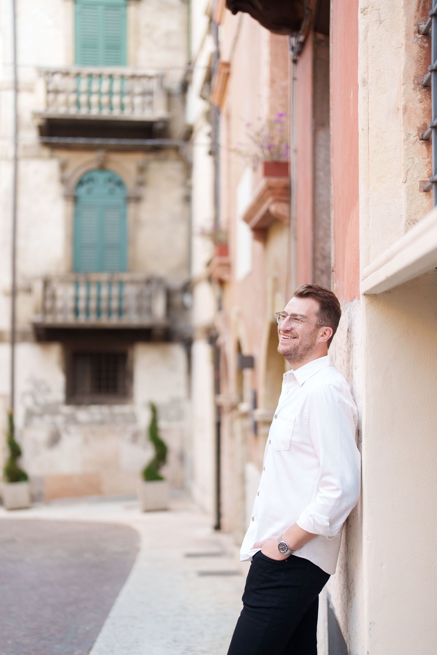 A person in a white dress shirt and black pants leans against a beige building wall on a cobblestone street.