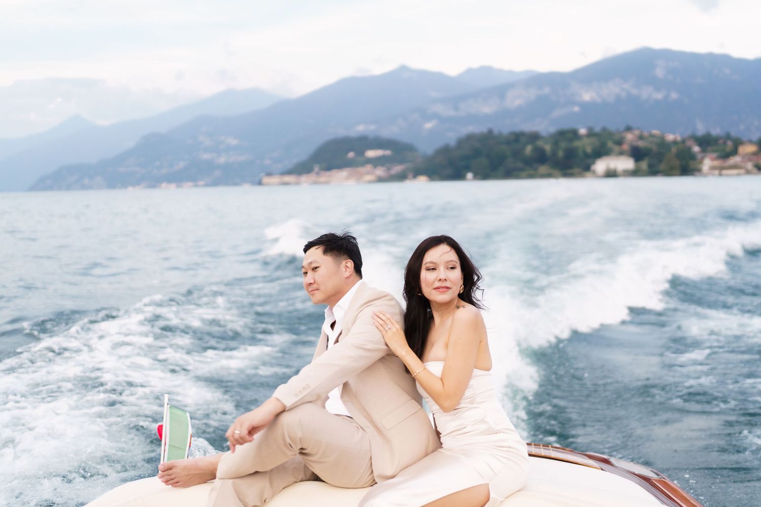A couple poses romantically on a boat against a scenic mountain backdrop on a sunny day.