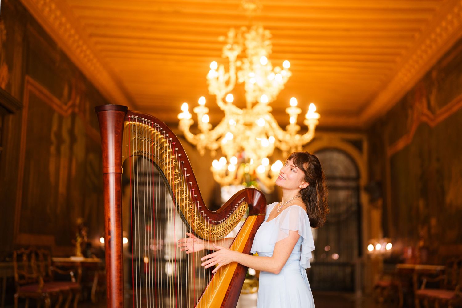 Elegant harpist performs in ornate ballroom with glowing crystal chandelier overhead.