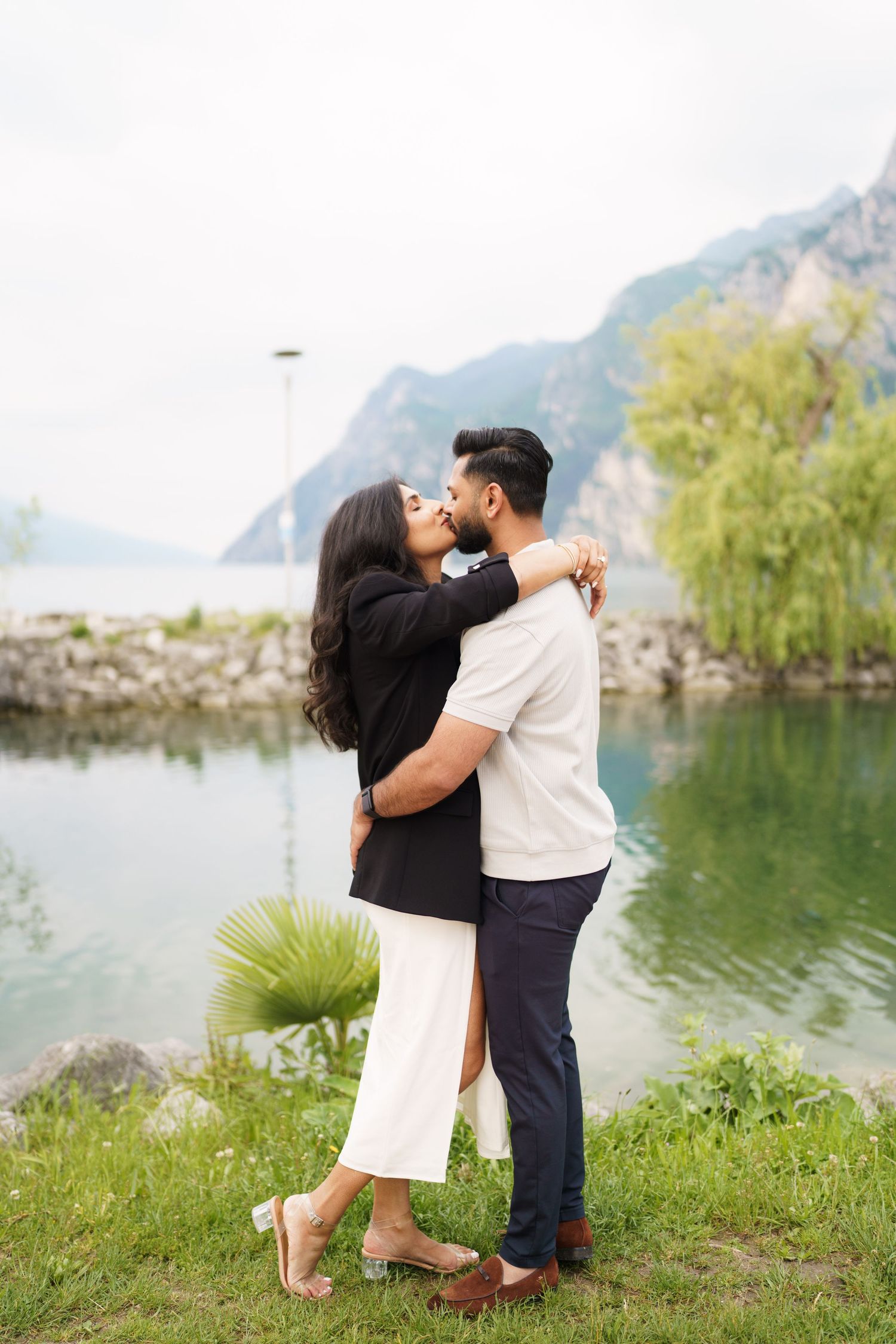 A romantic couple embraces and kisses by a serene mountain lake with rocky cliffs in the background.