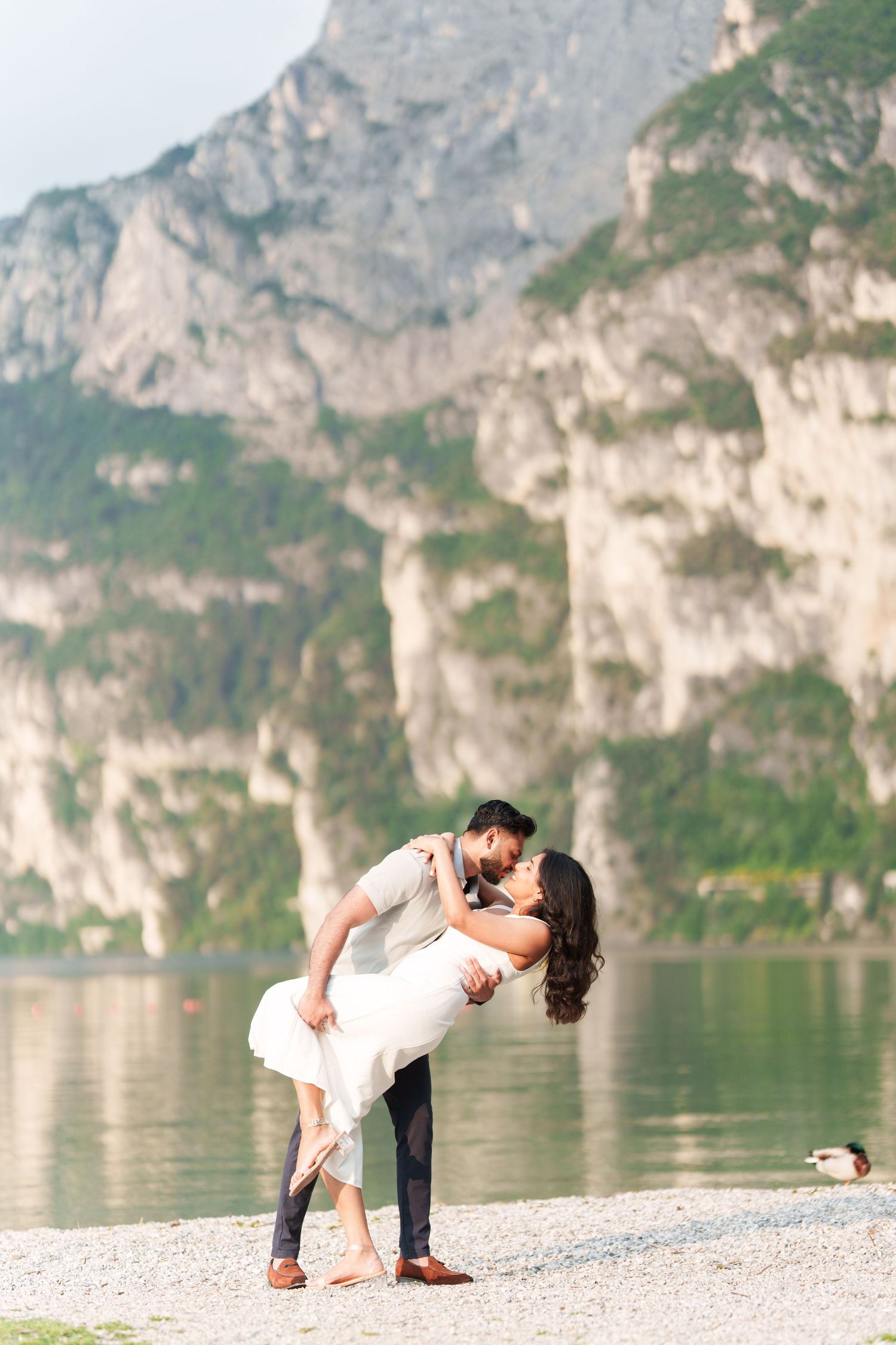 A romantic couple embraces on a rocky shore with dramatic mountain cliffs and a serene lake in the background.
