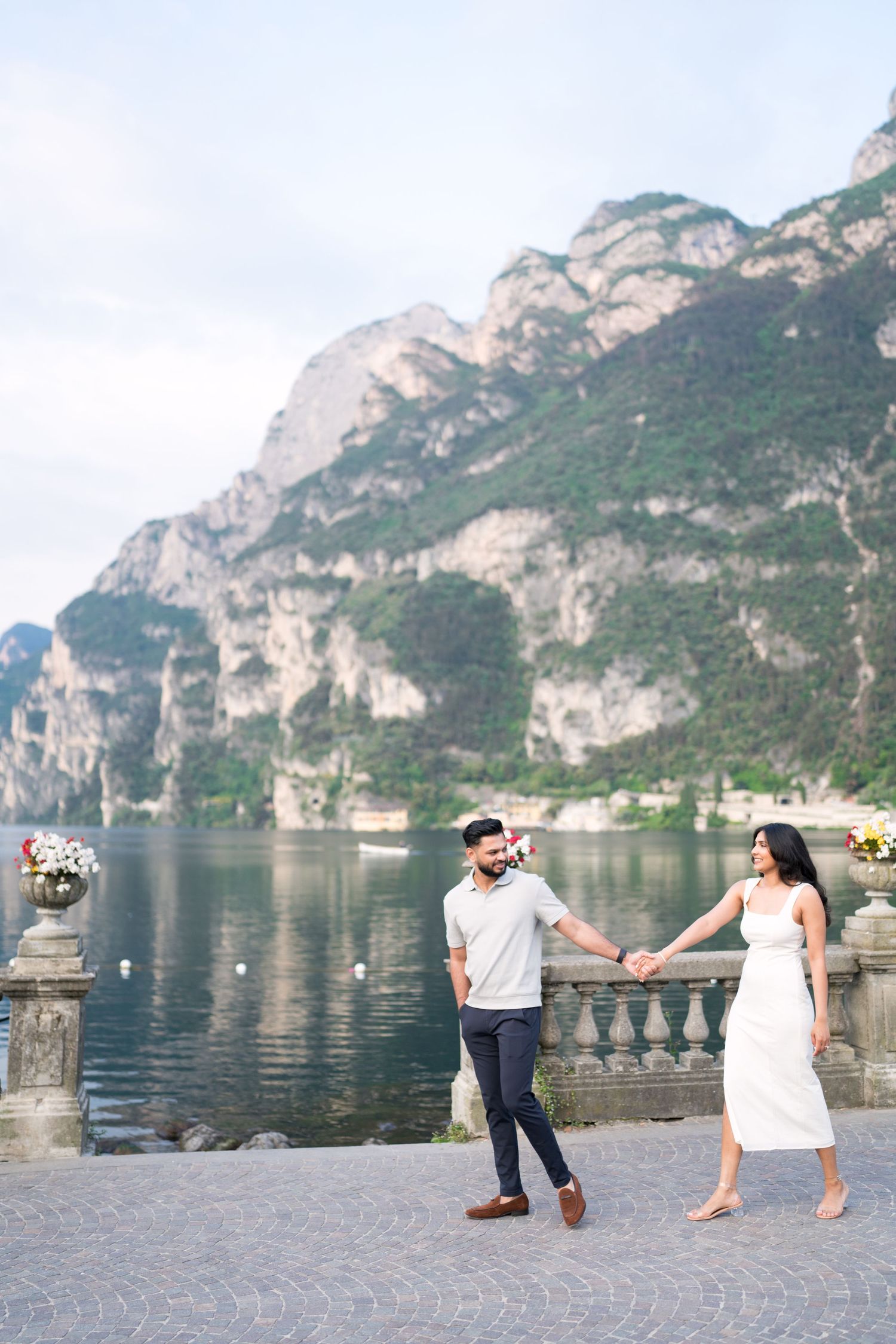 Couple enjoying romantic moments along a scenic lakeside promenade with dramatic mountain cliffs in the background.