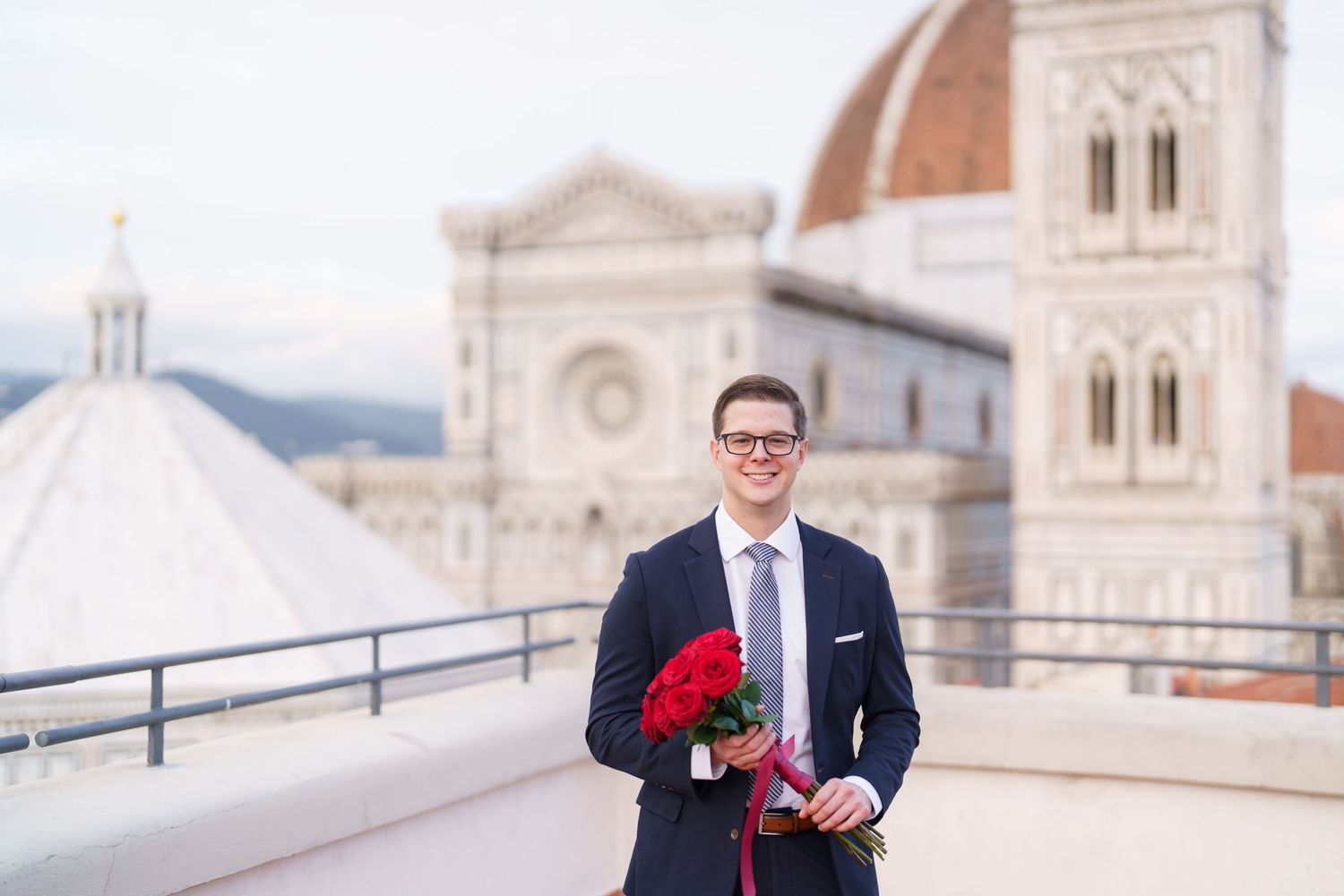 A person in a red sweater and suit stands on a rooftop terrace with historic European architecture in the background.