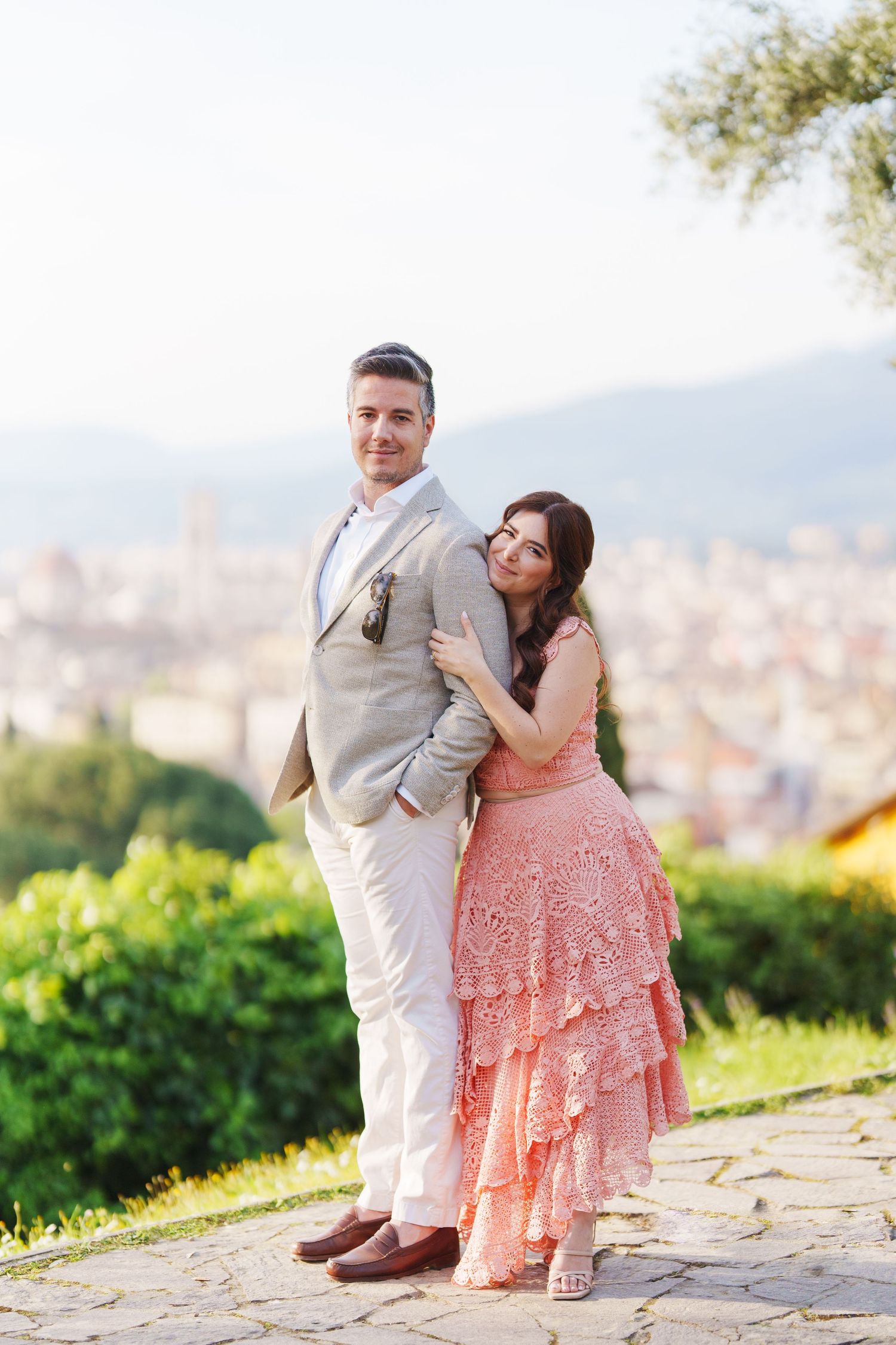 A couple poses together on a scenic overlook with a city vista behind them, wearing formal attire in beige and pink.