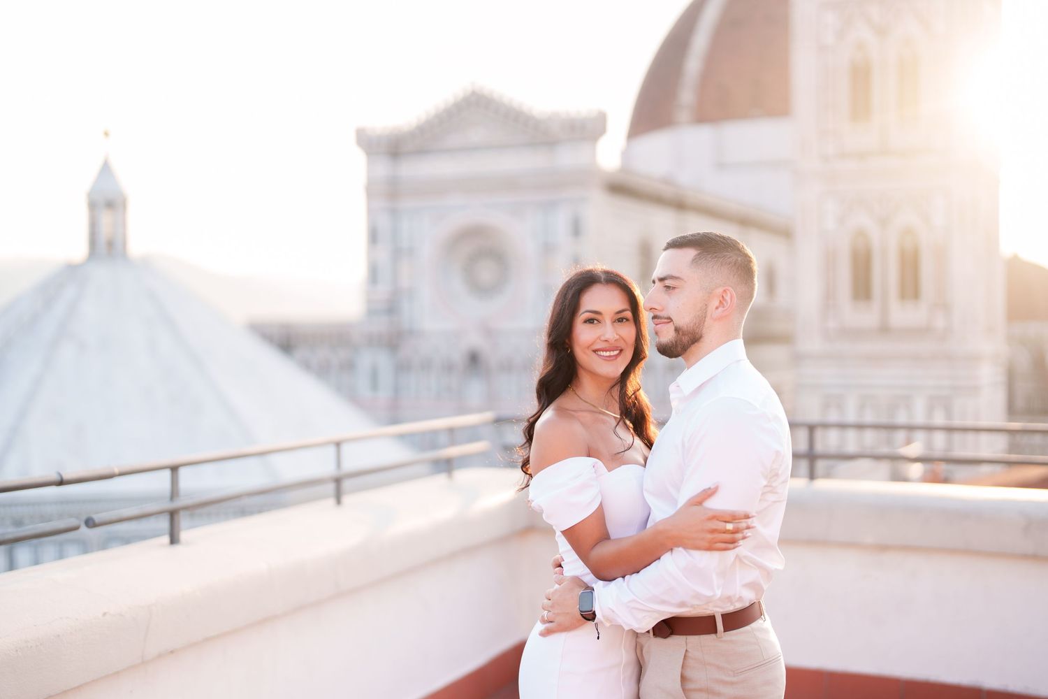 A couple in white outfits embraces on a rooftop terrace with a grand domed cathedral in the background.