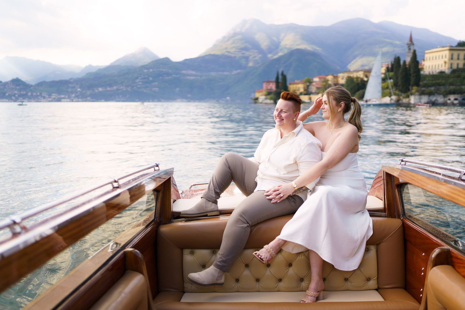 Couple sitting together on a classic wooden boat on Lake Como, Italy, with mountains and village in background.
