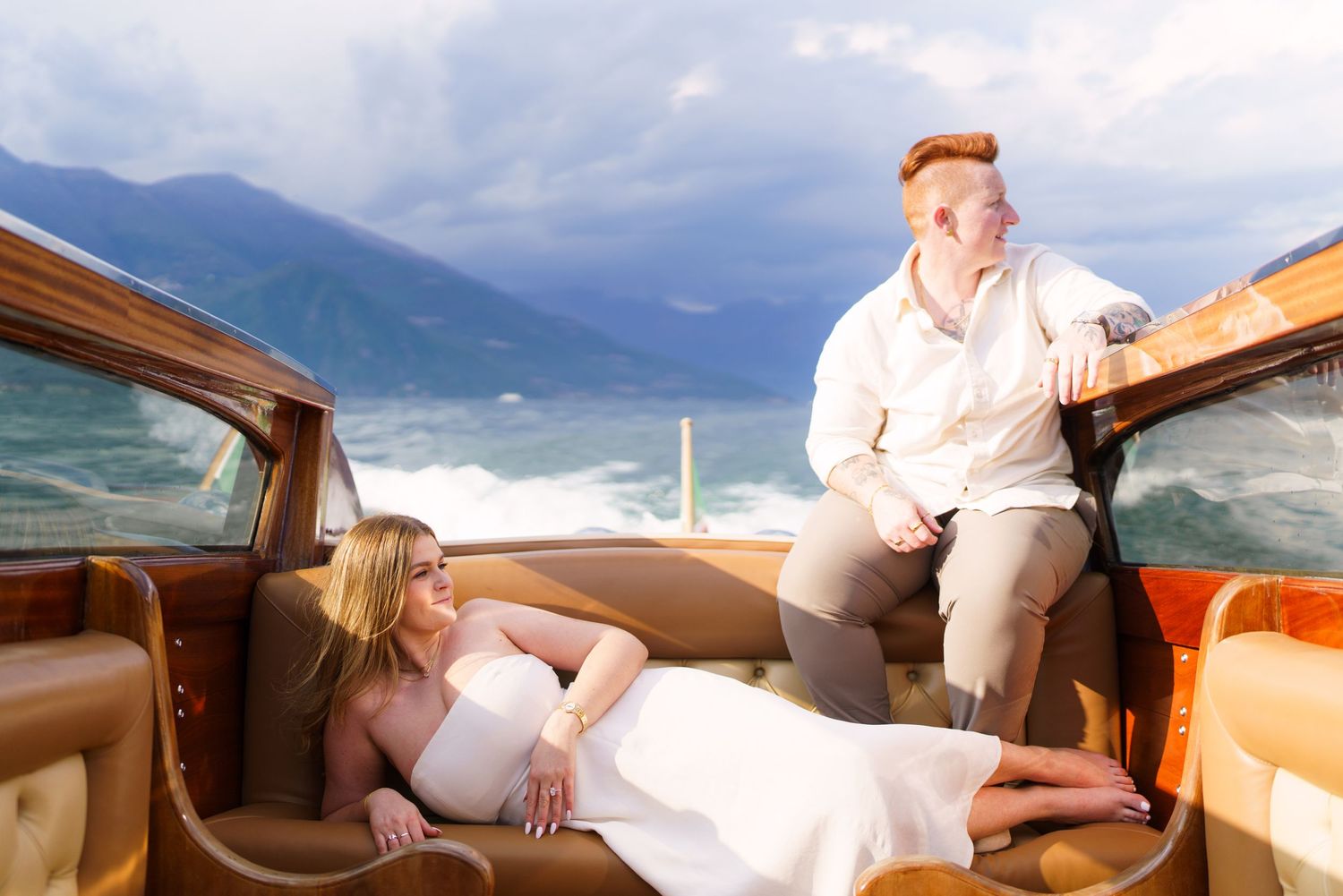 Couple posing on a luxury wooden boat with mountain lake backdrop, woman in white dress reclining, man seated casually.