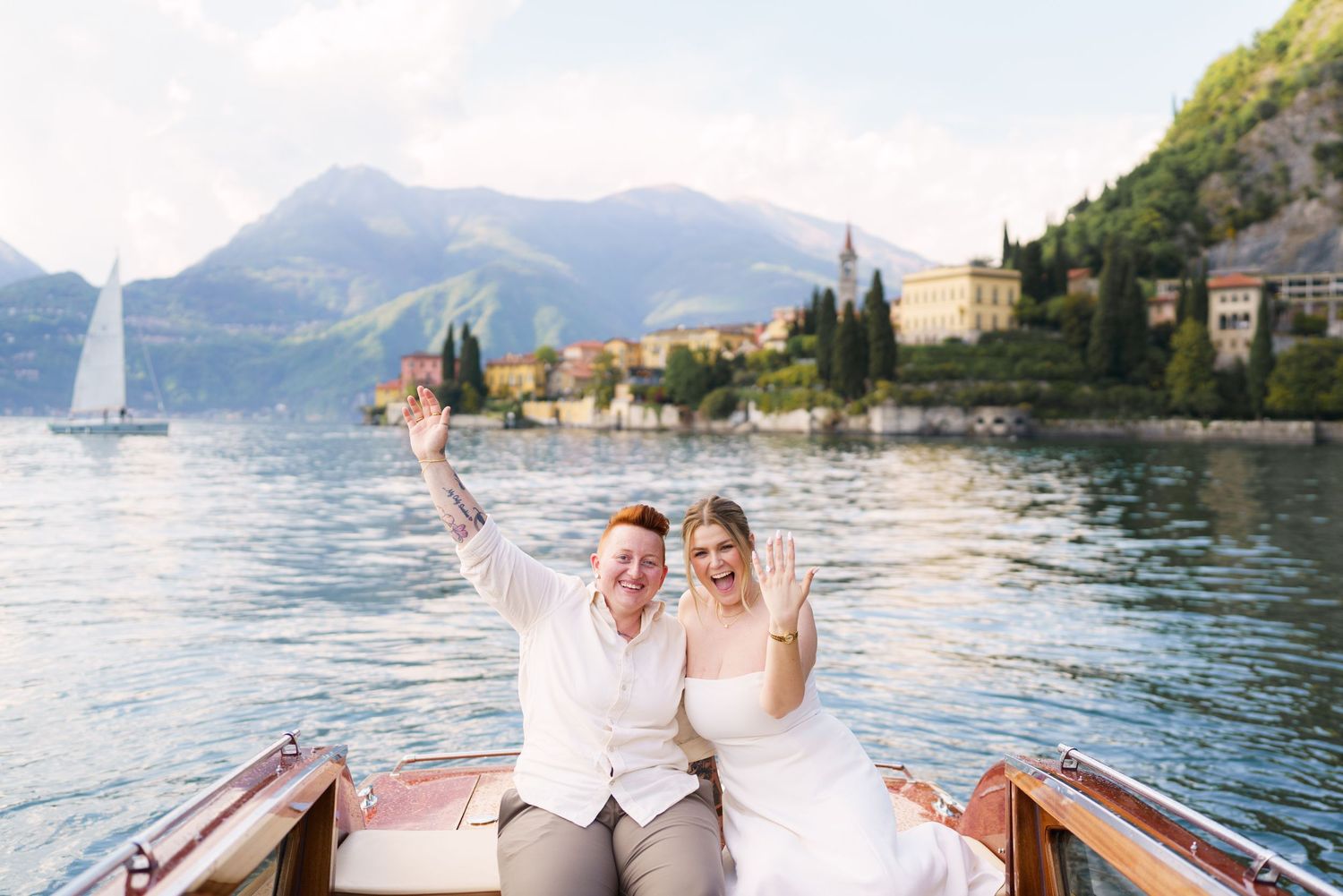 Joyful newlyweds celebrate on a wooden boat on Lake Como, Italy, with mountains and a sailboat in the background.
