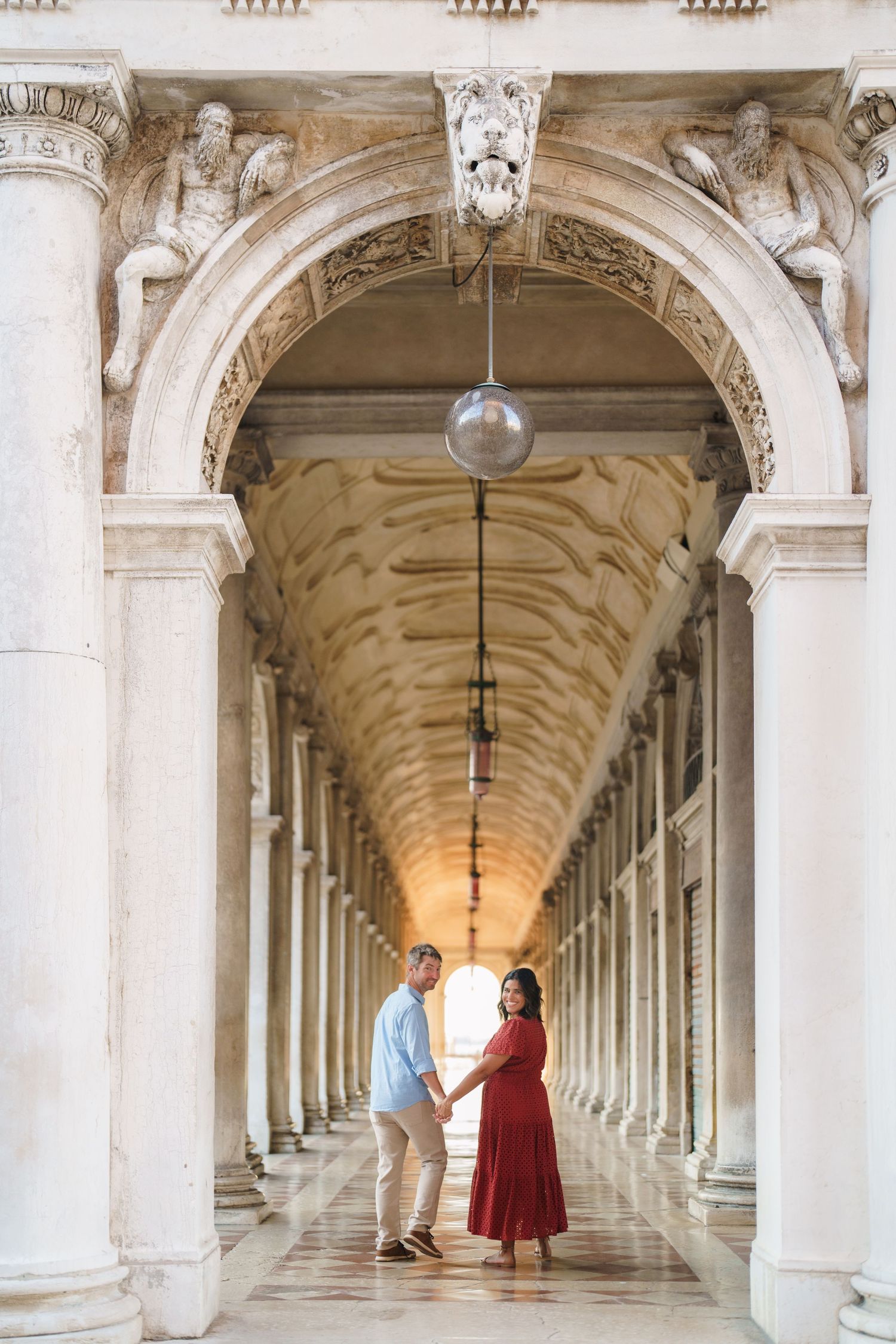 Couple holding hands under ornate marble arches of a classical European colonnade, woman in red dress, man in blue shirt.