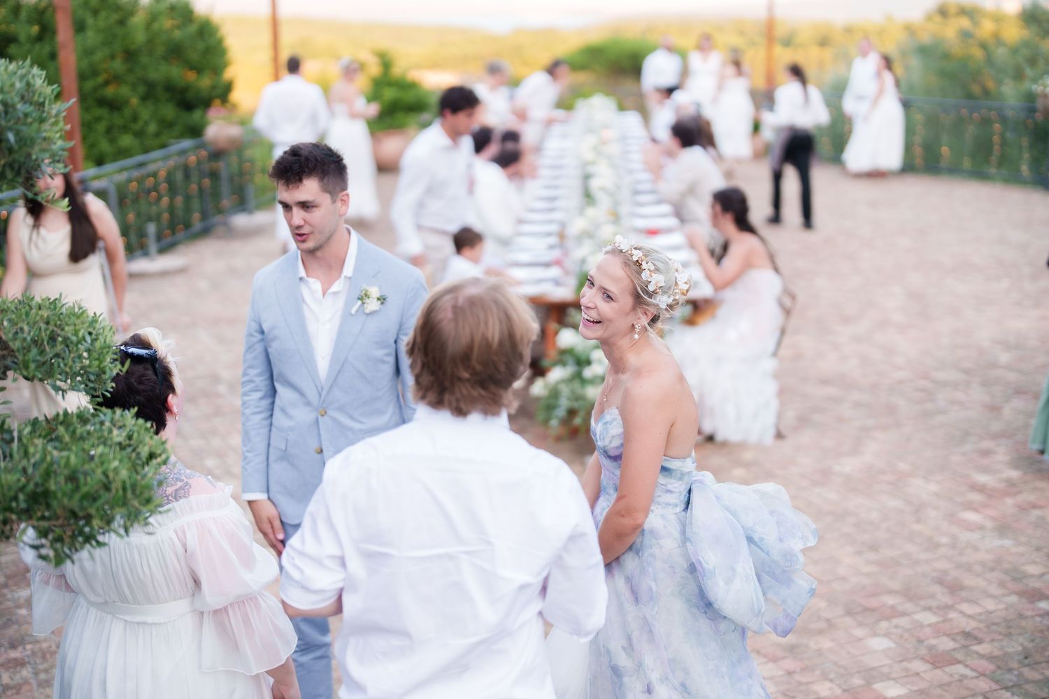Elegant outdoor wedding reception with guests in white and blue attire mingling on a sunlit terrace.