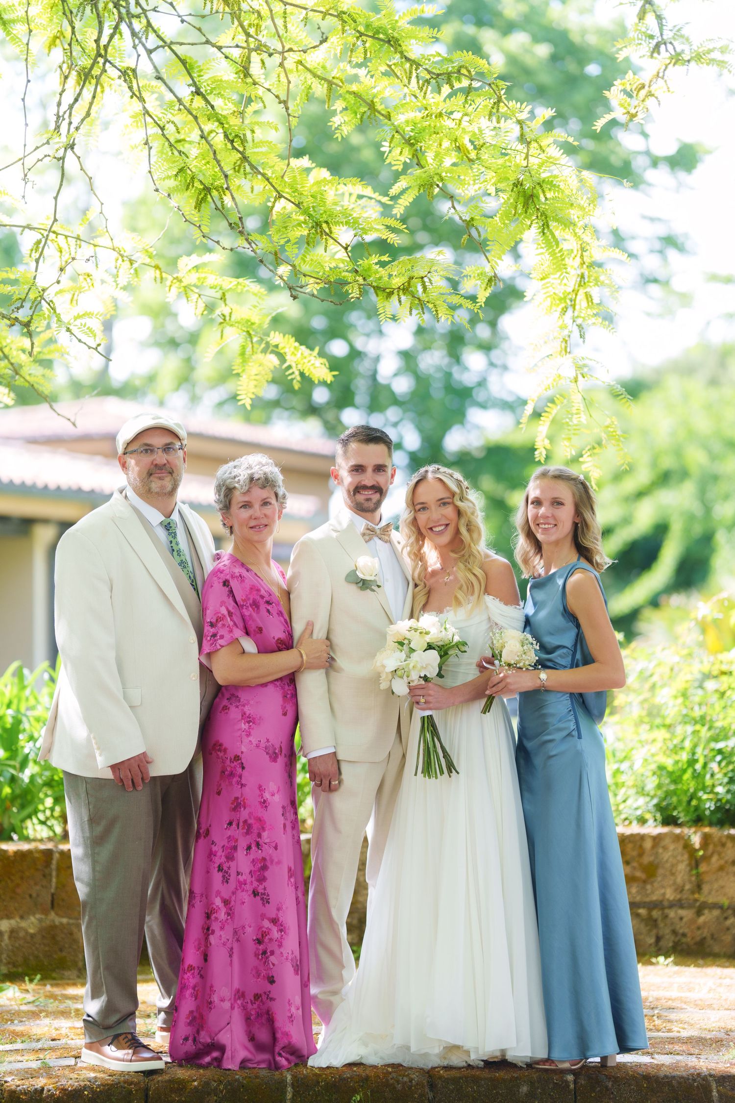 Wedding party posing outdoors with bride in white gown, groom in tan suit, and guests in pink and blue dresses.