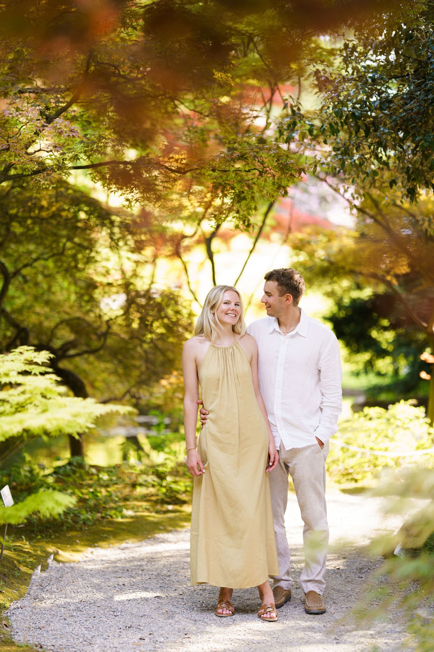 Couple walking hand in hand on a garden path surrounded by lush green and autumn-colored trees.
