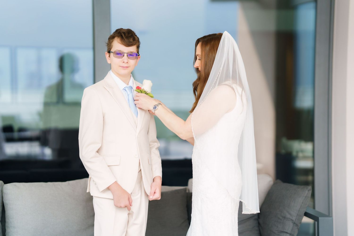 Bride in white veil pins boutonniere on groom in beige suit with purple glasses at wedding venue.