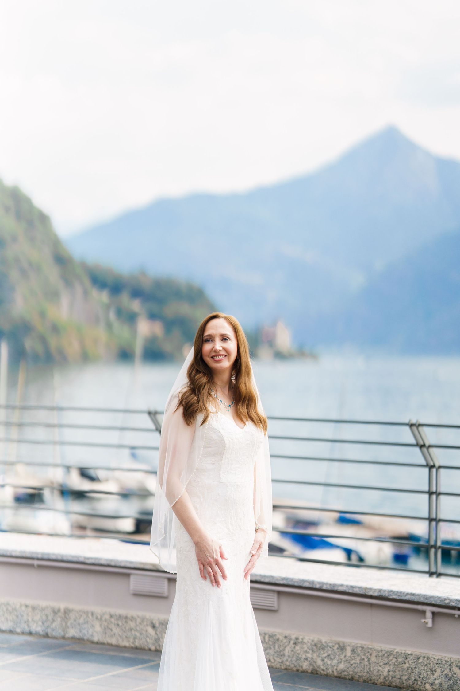 Bride in white gown stands on lakeside terrace with mountain backdrop, smiling elegantly on her wedding day.