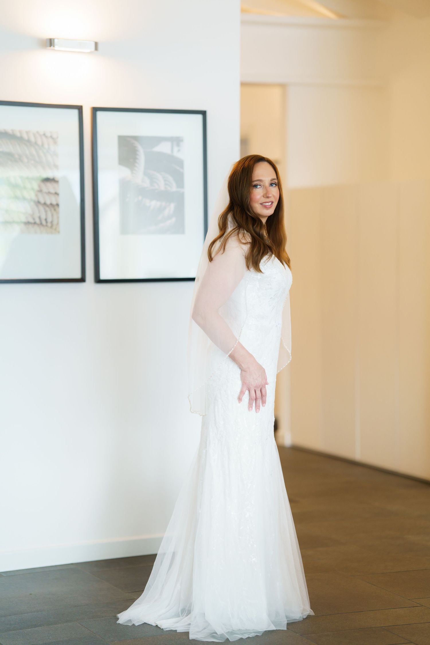 A smiling bride in a strapless white tulle wedding gown poses elegantly in a bright room with framed artwork.