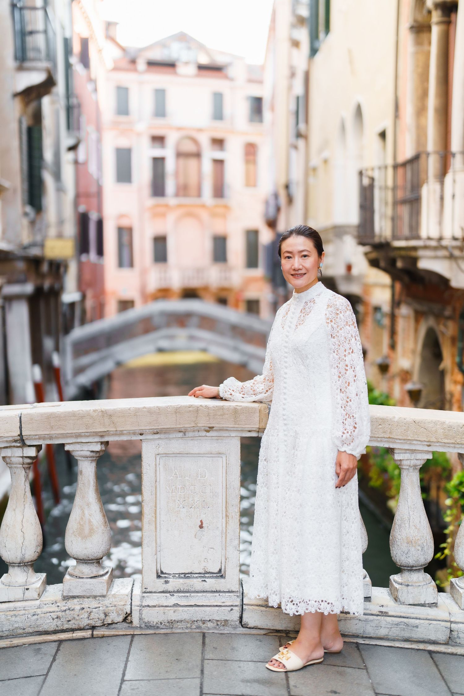 Woman in white lace dress posing on a Venice canal bridge with historic buildings in the background.