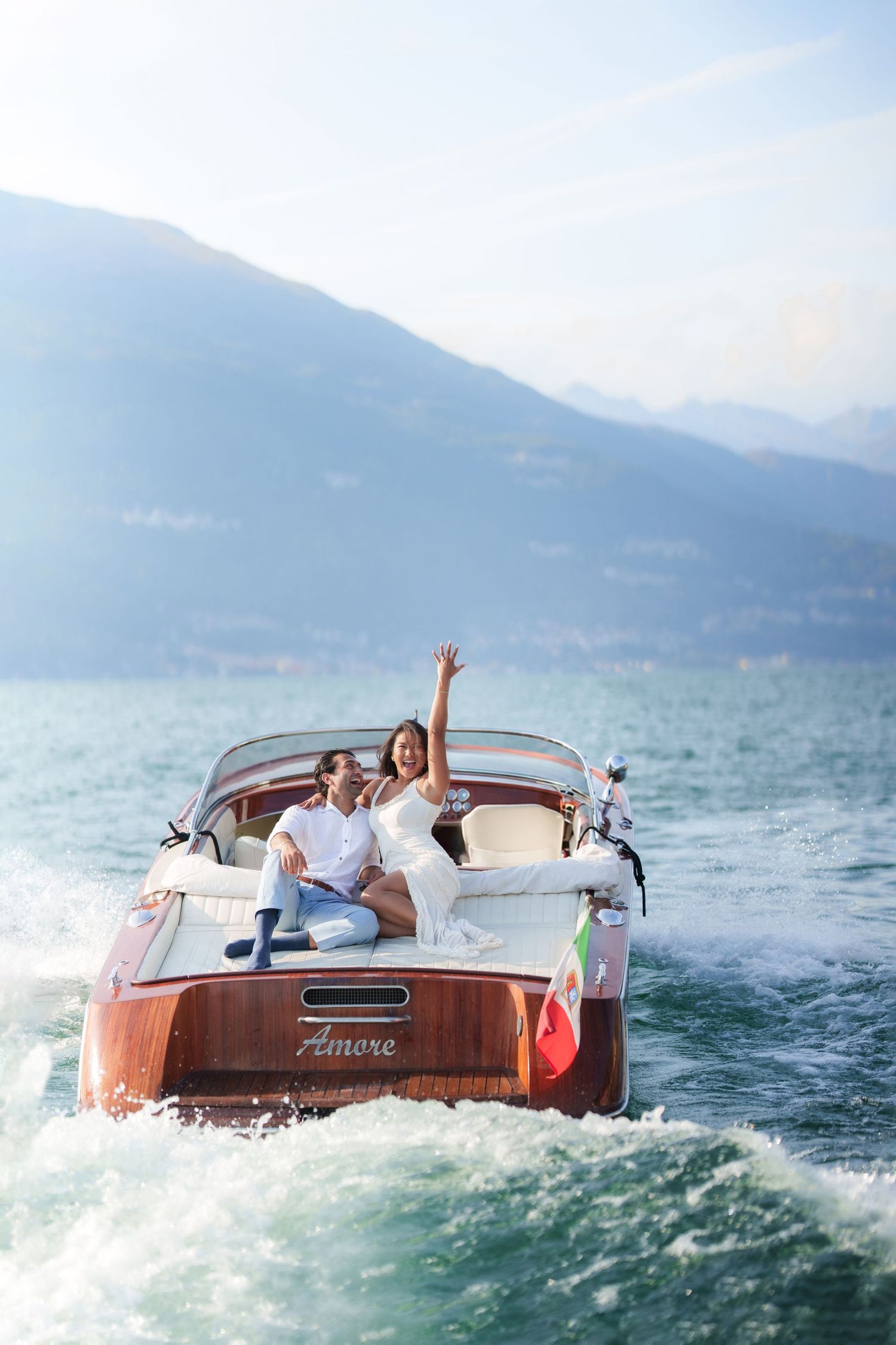 Couple celebrating on a wooden Amore boat on a scenic lake with mountain backdrop, woman waving joyfully.