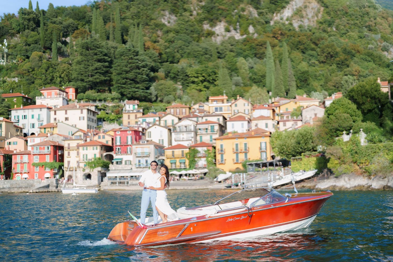 Couple standing on a red speedboat on Lake Como with colorful Italian village buildings and green hills in background.