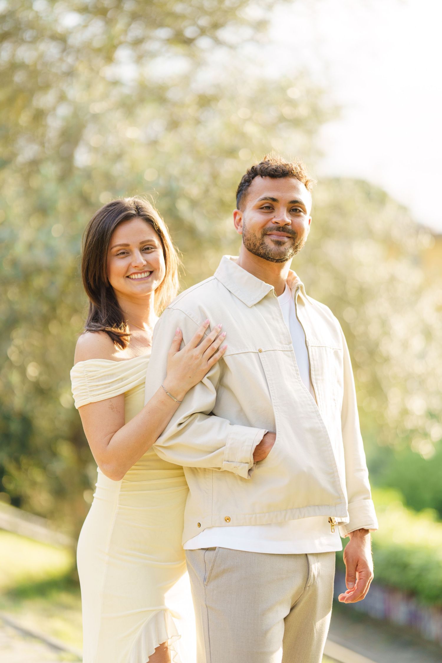 Happy couple in beige outfits posing outdoors in sunlit park, woman in yellow dress embracing man in cream jacket.