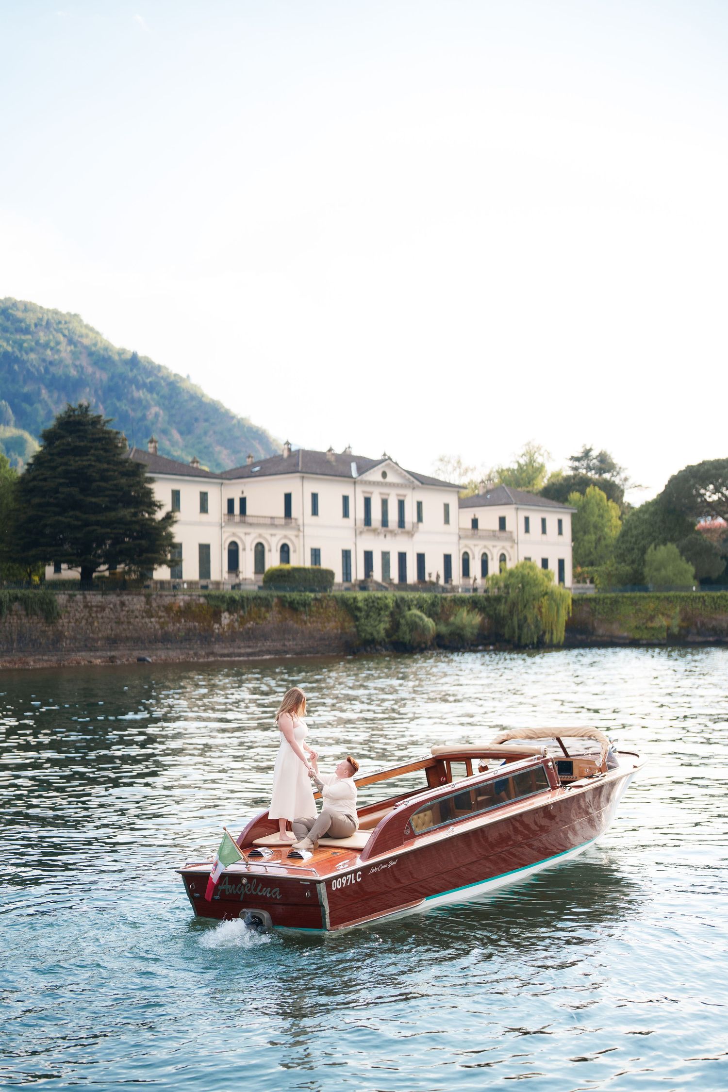 Couple in wedding attire riding a classic wooden boat on Lake Como with elegant Italian villa in background.