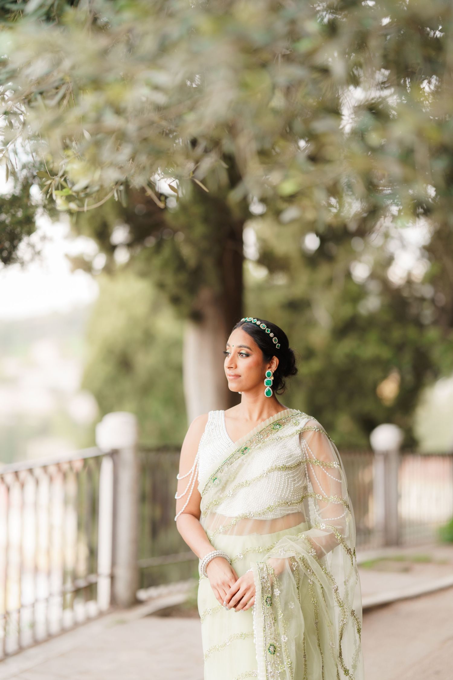 A person in a traditional mint green saree poses elegantly against a blurred outdoor backdrop with white railings.