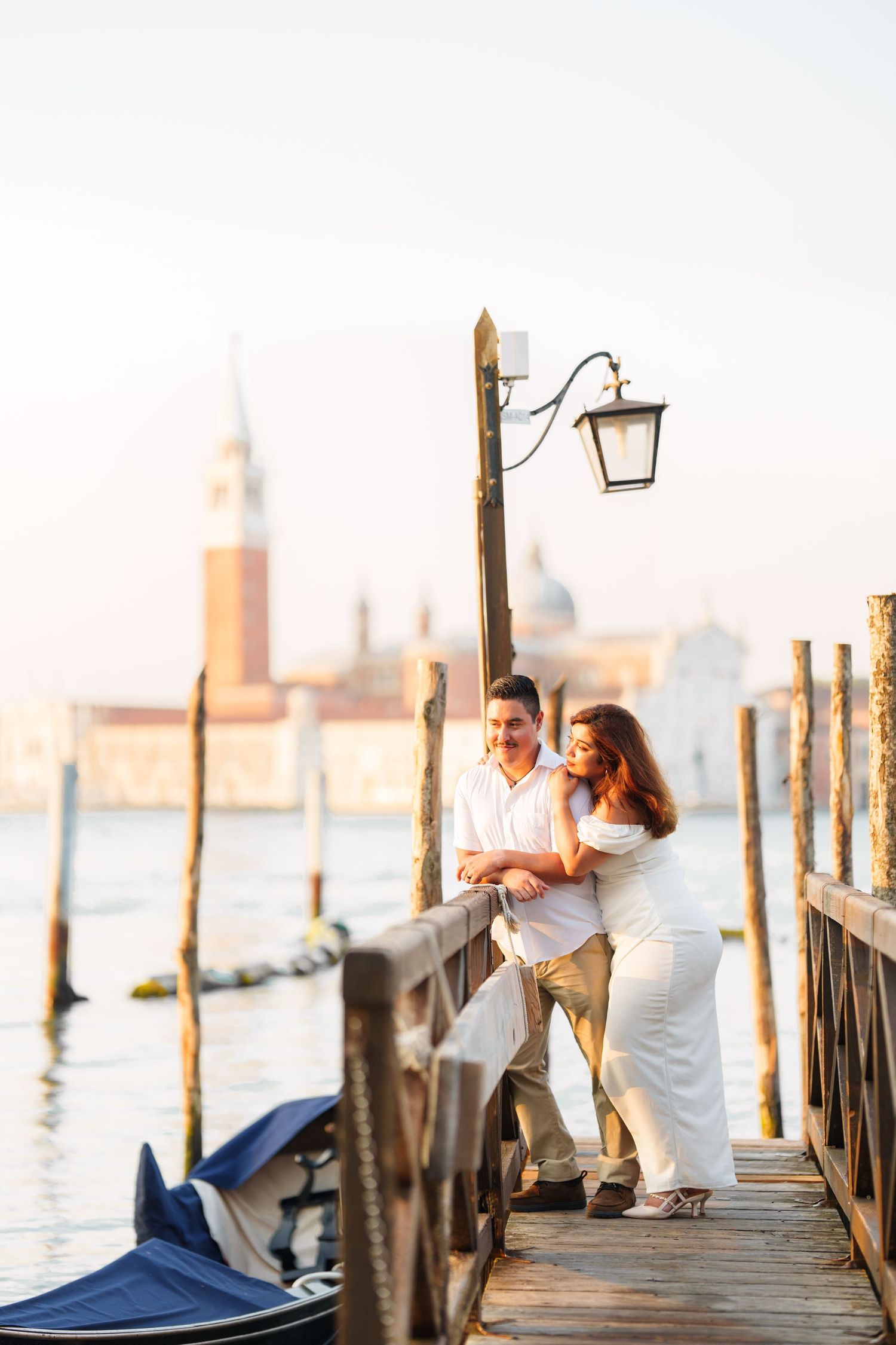 Romantic couple embraces on a wooden pier overlooking Venice's Grand Canal at sunset with gondolas in the background.