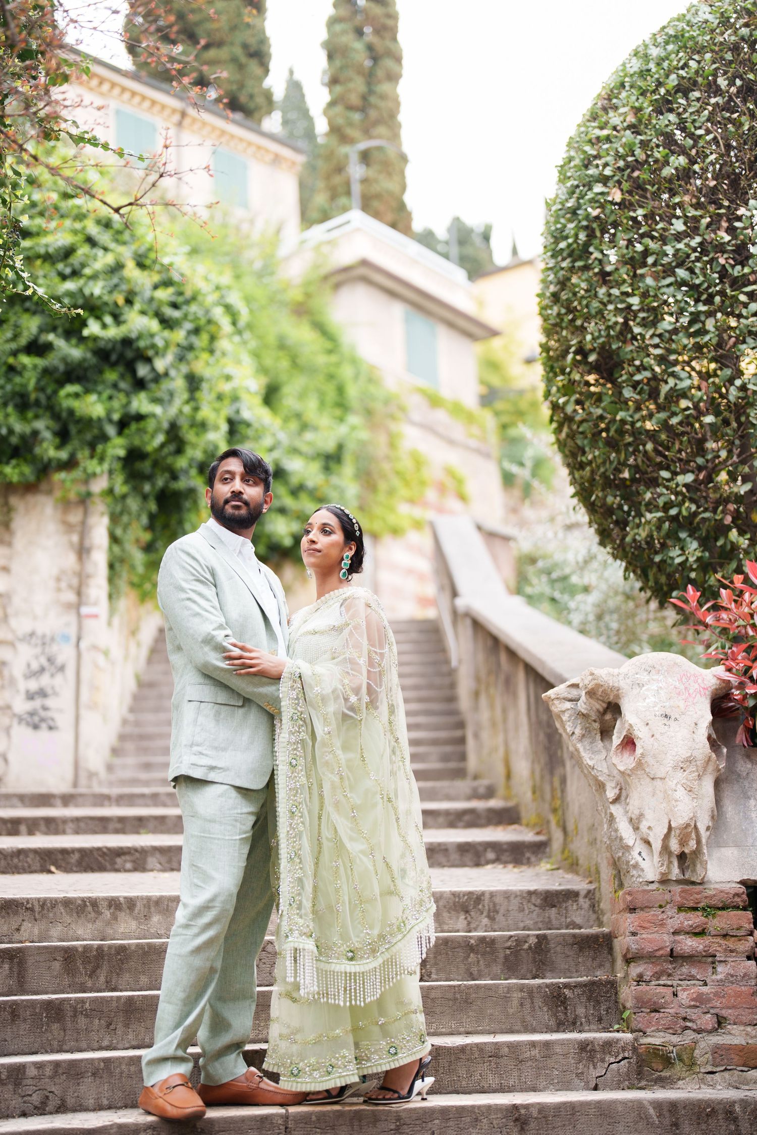 A couple in elegant mint green traditional South Asian attire pose together on stone steps in a garden setting.