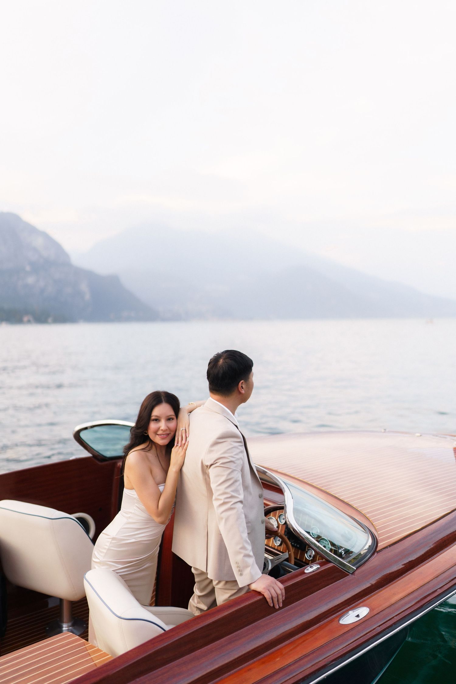 A couple enjoys a romantic boat ride in a vintage wooden speedboat on a misty lake surrounded by mountains.