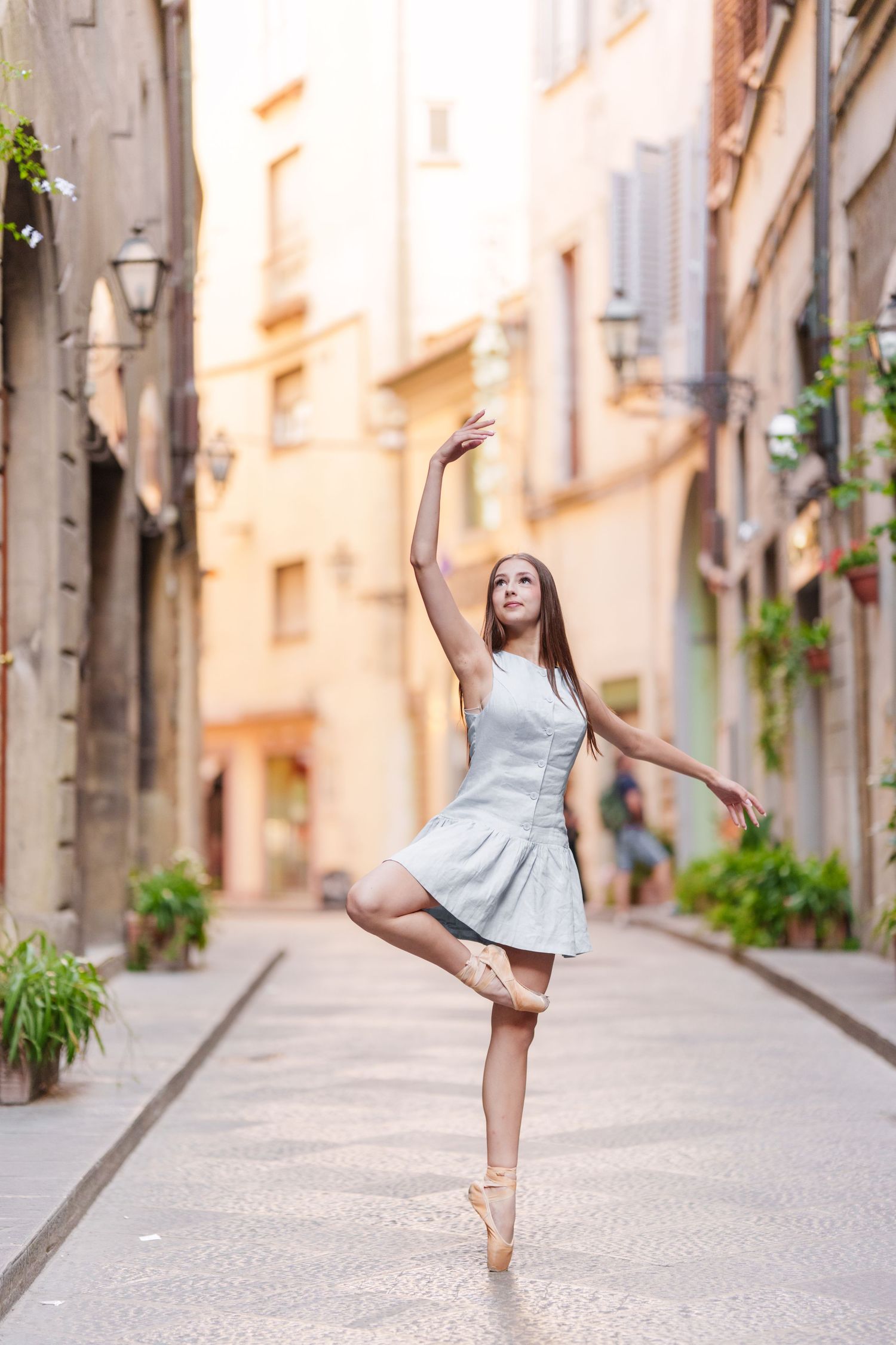 Ballet dancer in light dress performs graceful pose en pointe in charming European alley with potted plants.