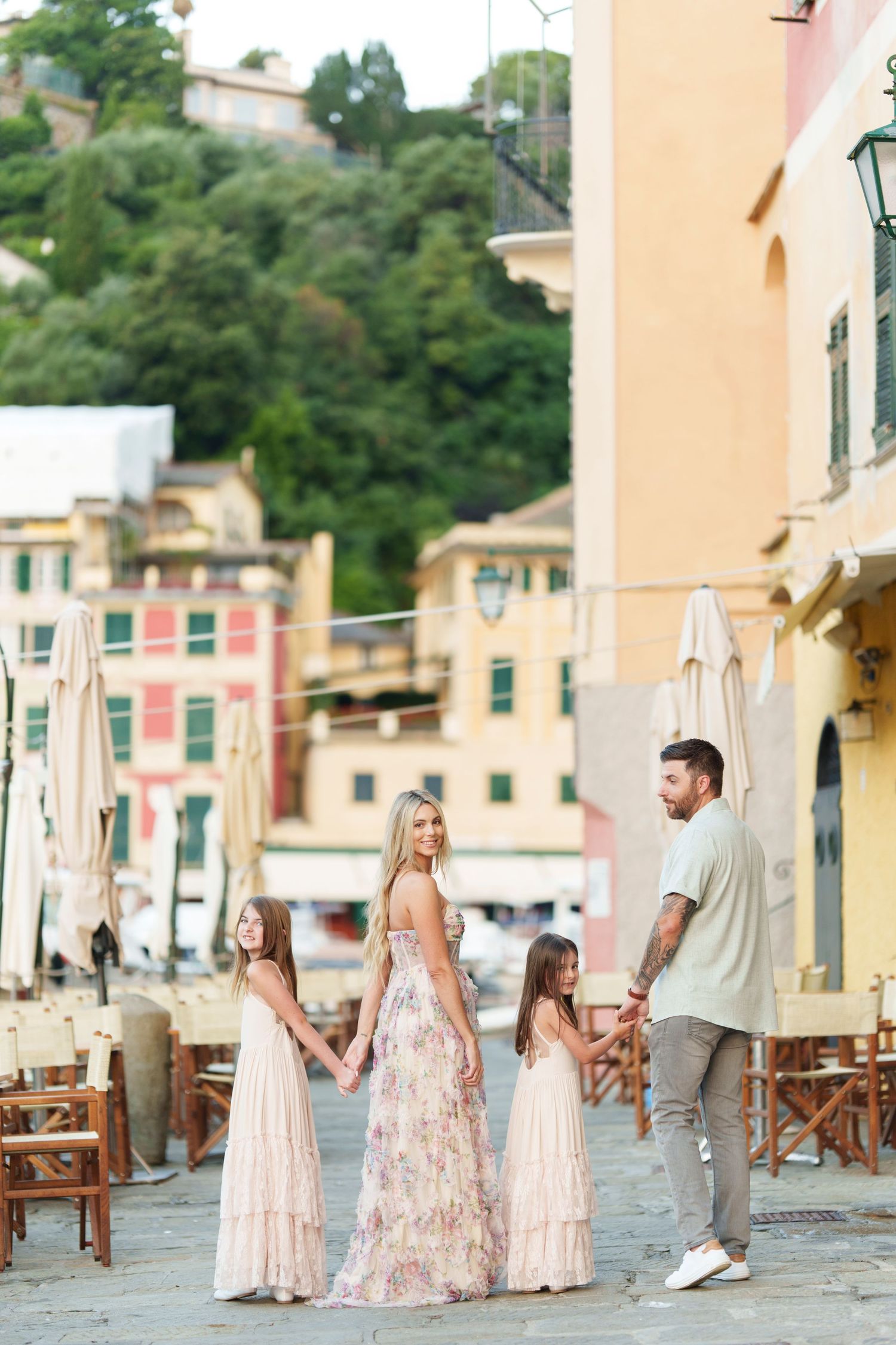 A family in coordinated pink and beige outfits walks along a charming Mediterranean coastal street with pastel buildings.
