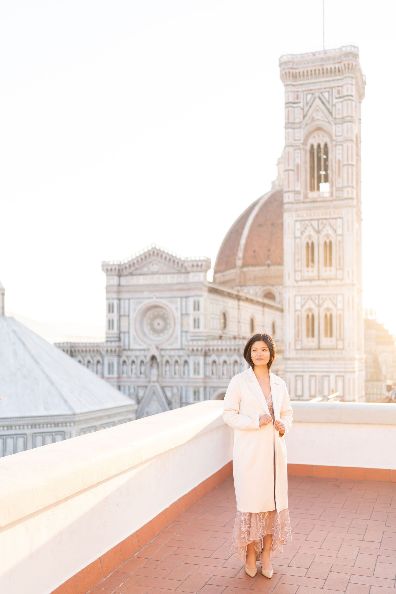 A person in a white dress stands on a terrace overlooking the iconic Duomo cathedral in Florence, Italy at sunset.
