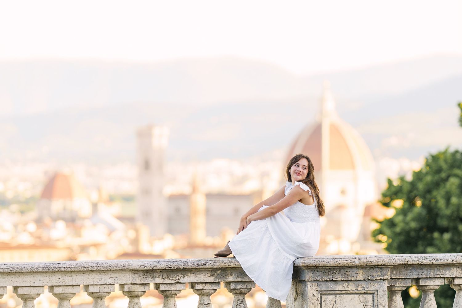 A woman in a flowing white dress poses gracefully on a stone balcony overlooking Florence, Italy at sunset.