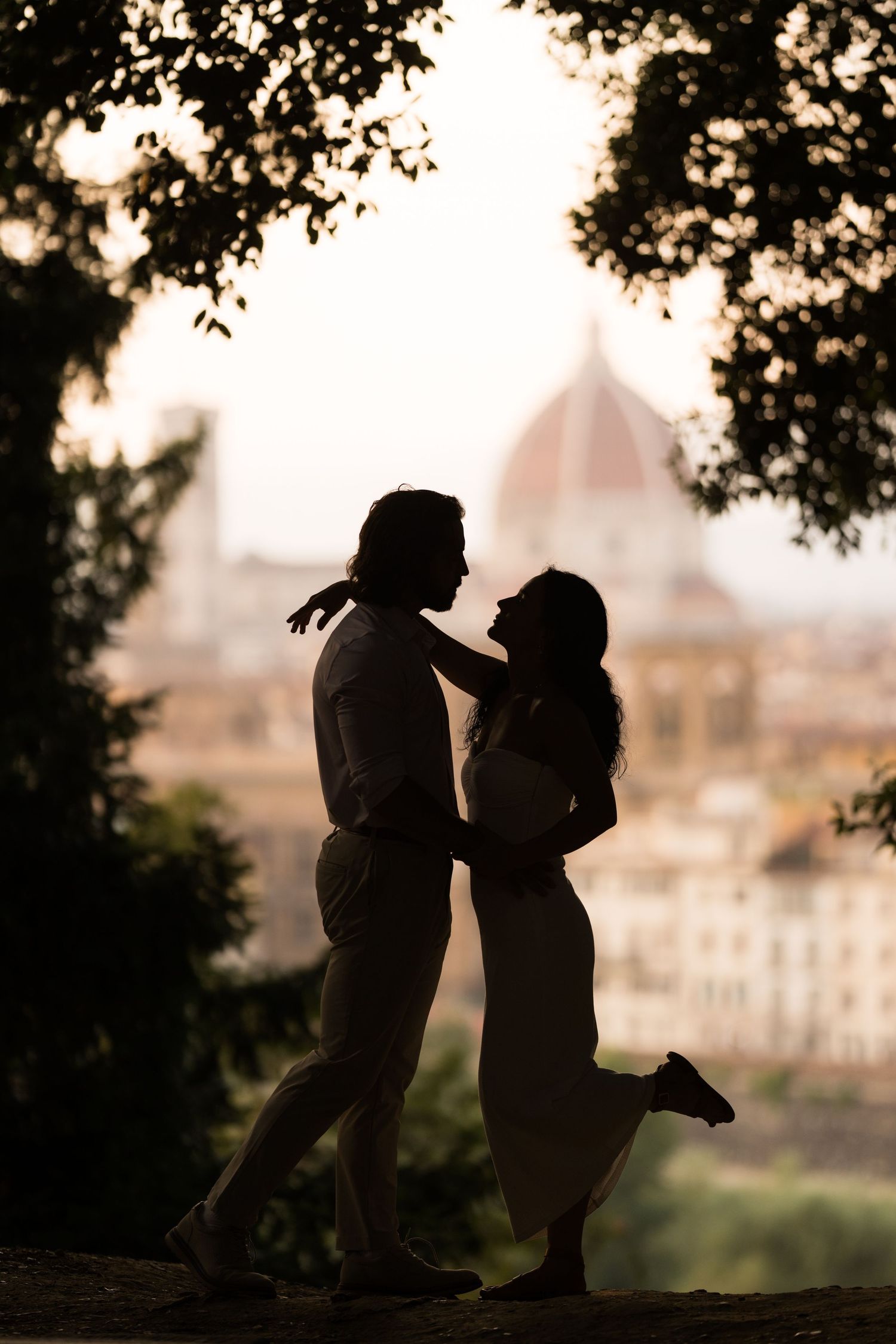 Silhouetted couple shares romantic moment against blurred cityscape backdrop at sunset.
