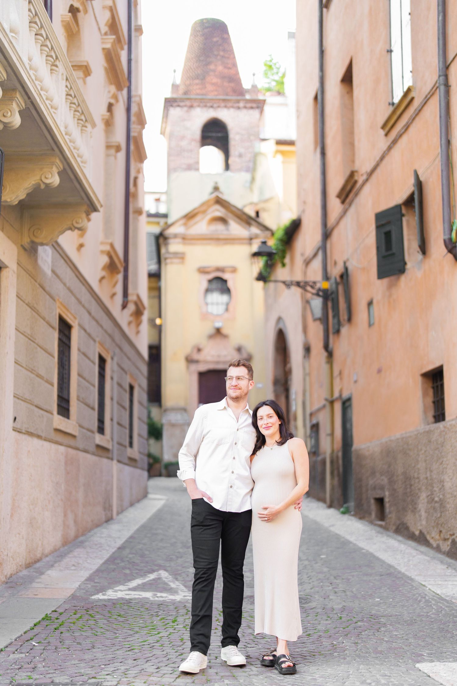 A romantic couple strolls down a charming cobblestone alley in an old European town with peach-colored buildings.