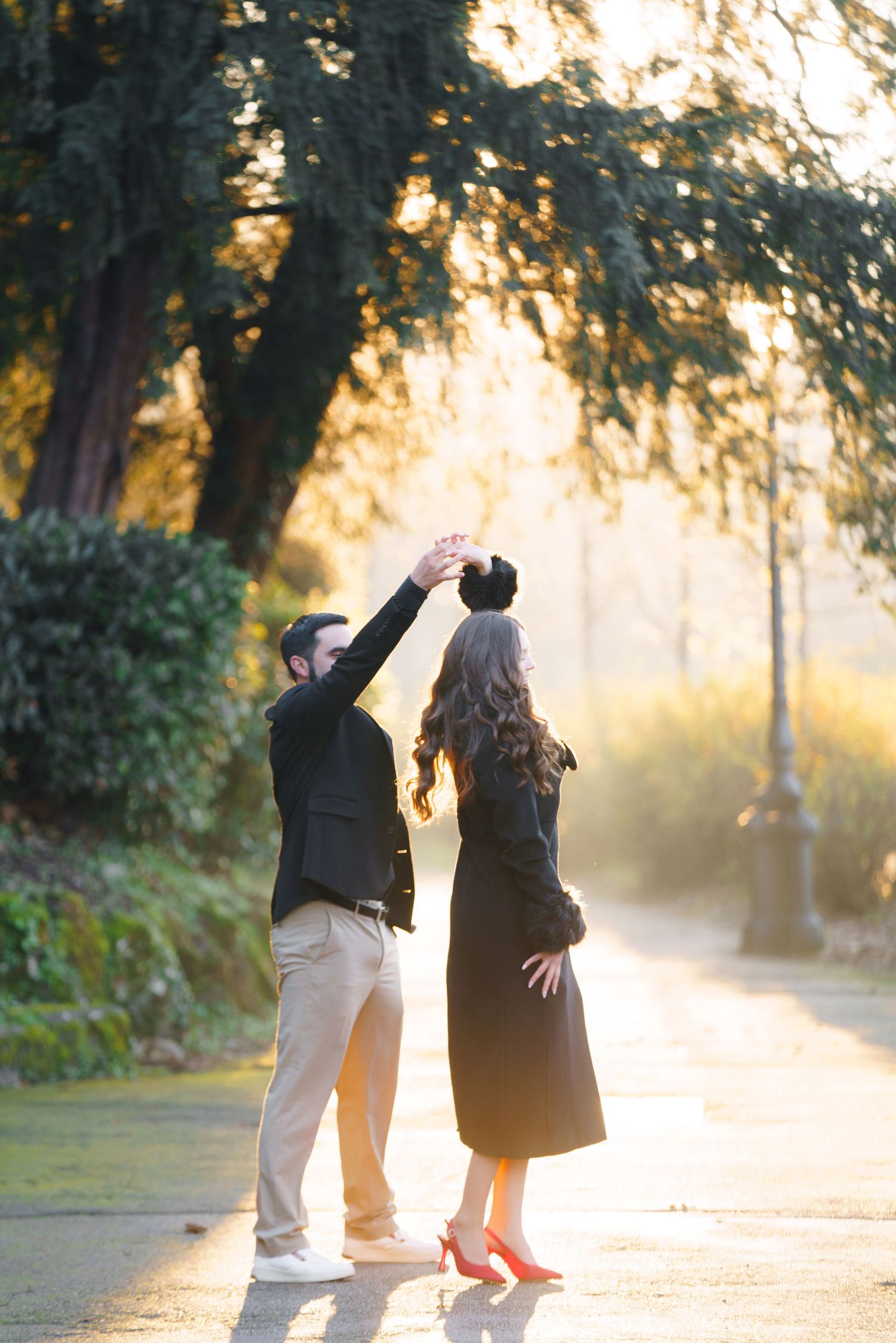 A couple walks together on a path at sunset with golden sunlight streaming through trees in a romantic setting.