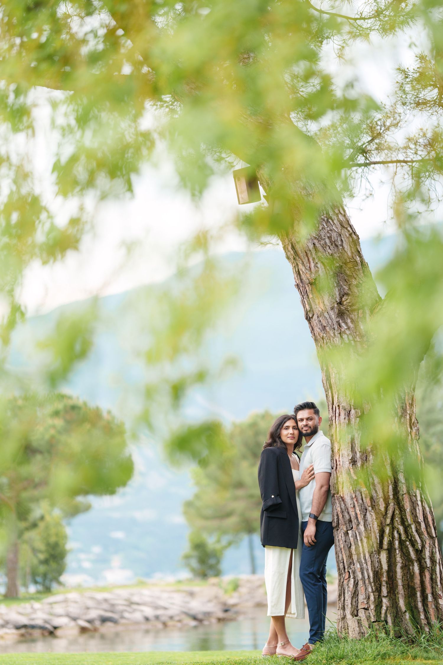 A couple shares a romantic moment among trees by a lakeside on a bright sunny day.