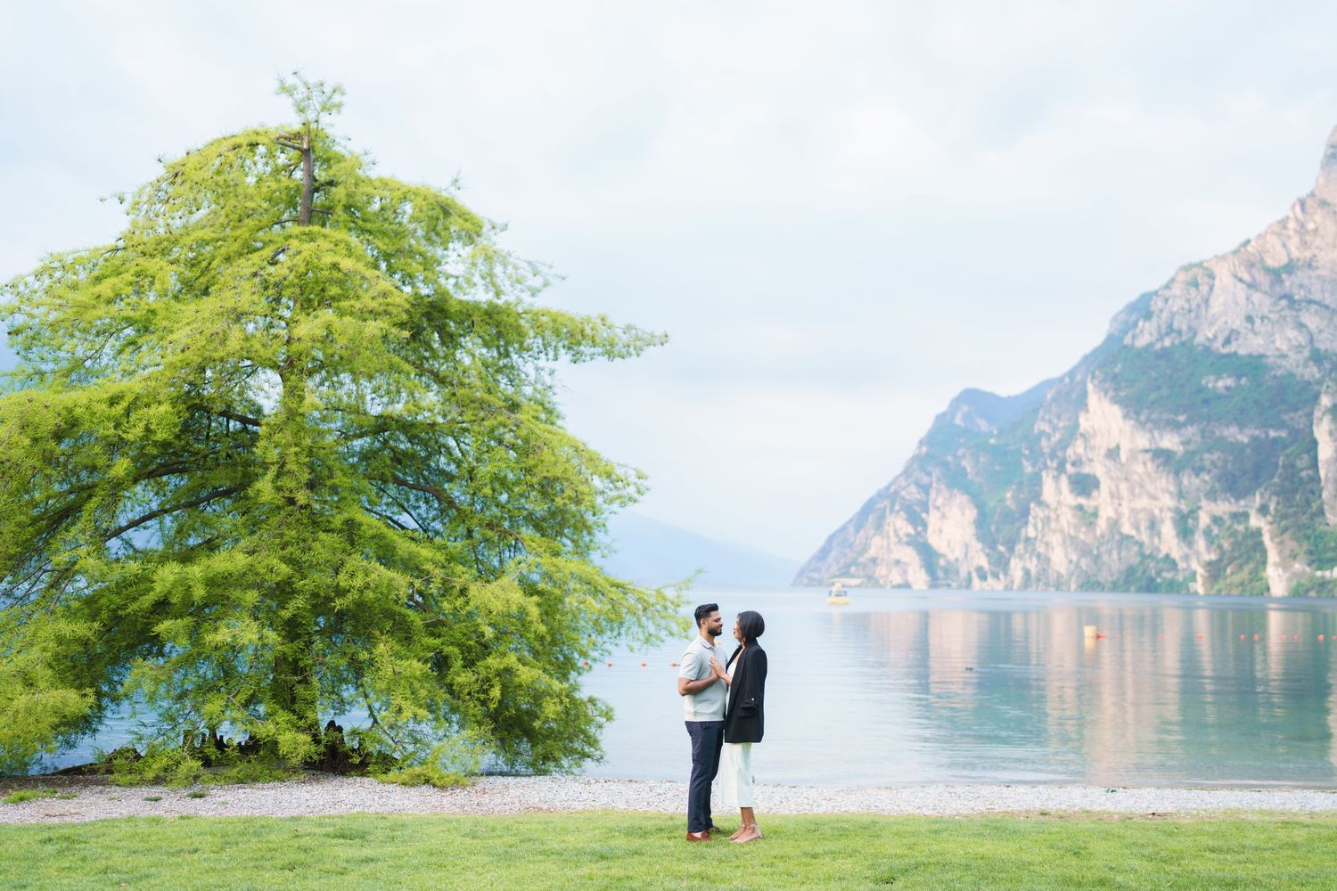 A couple embraces by a scenic mountain lake with a lush green tree in the foreground.