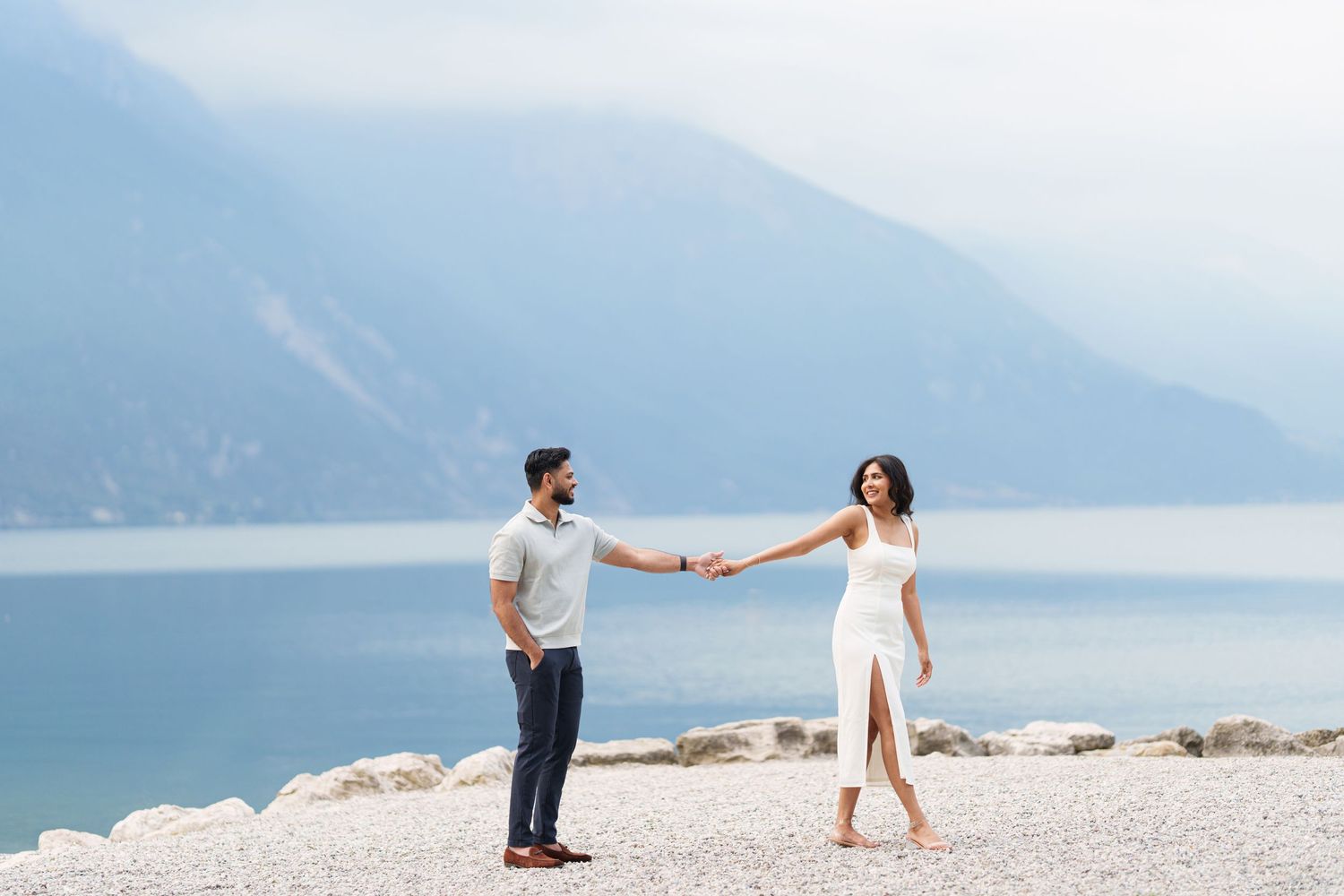 Two people hold hands while standing on a rocky beach with misty mountains and a lake in the background.