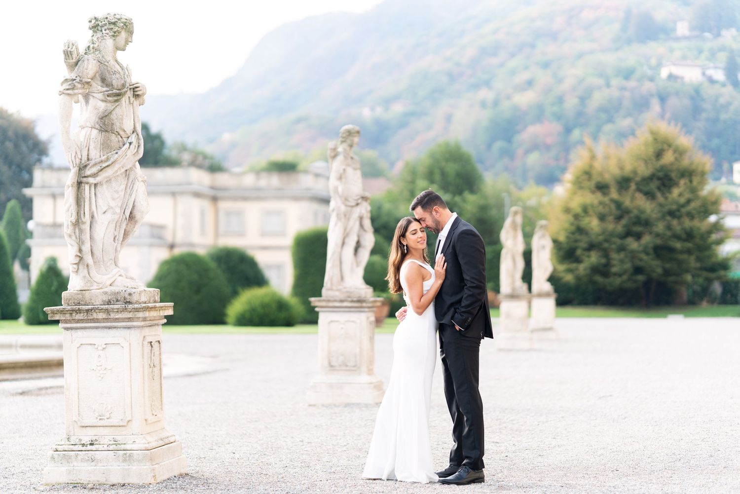 A romantic wedding couple poses near ornate stone statues in an elegant garden setting with mountain views.