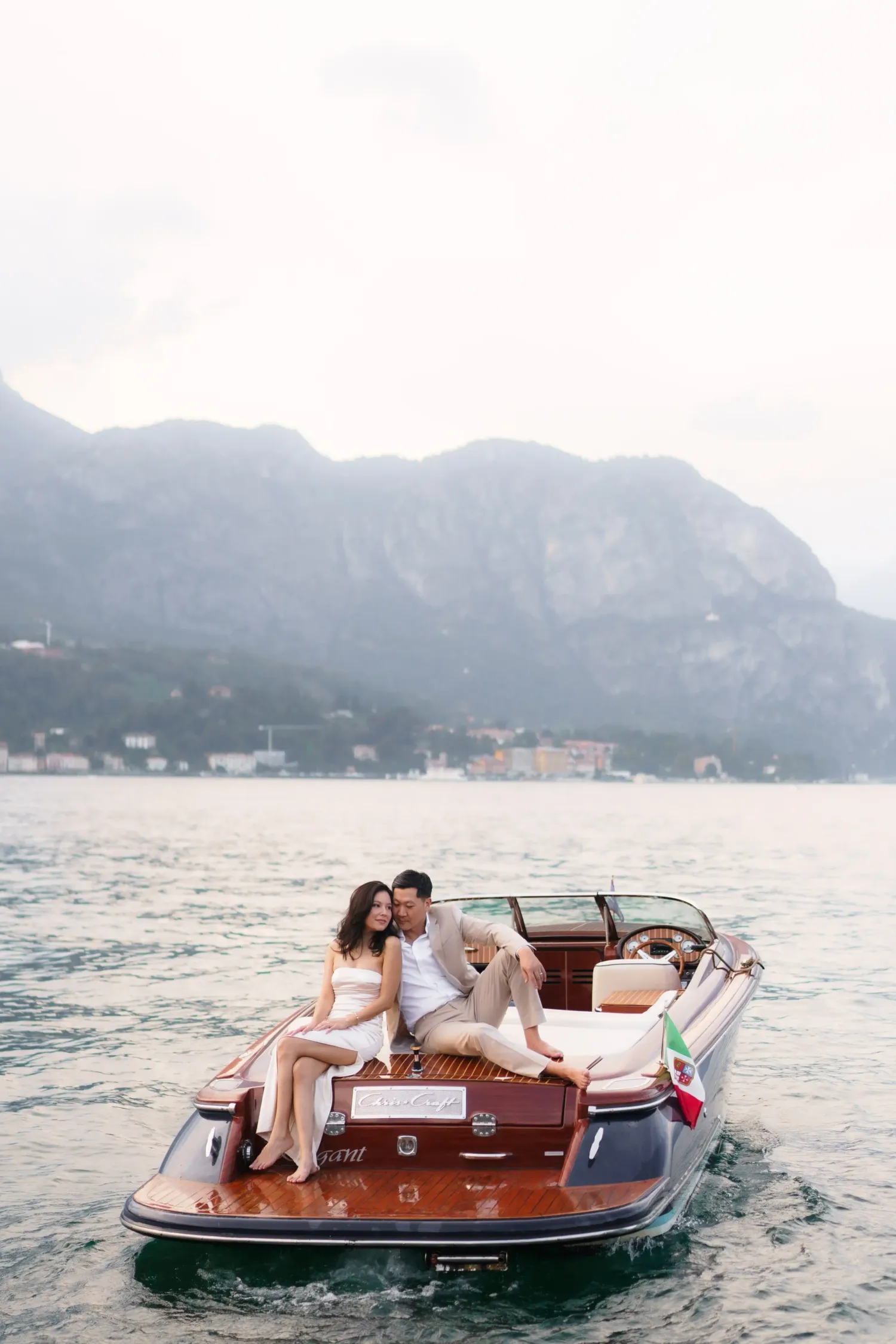 Romantic couple enjoying a scenic boat ride across a misty lake surrounded by mountains in a classic wooden vessel.