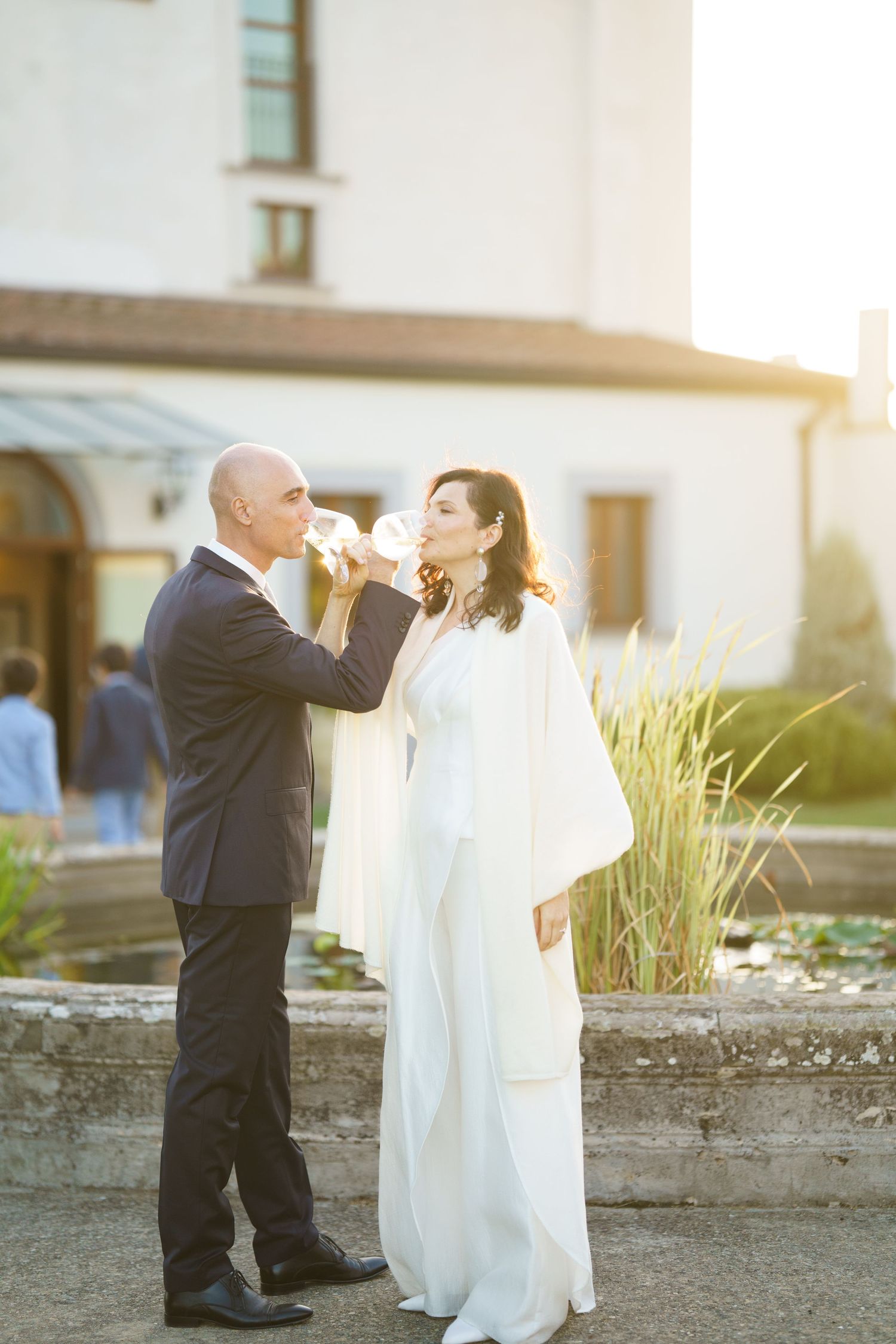 A couple shares a playful moment with wine glasses at their outdoor wedding during golden hour.