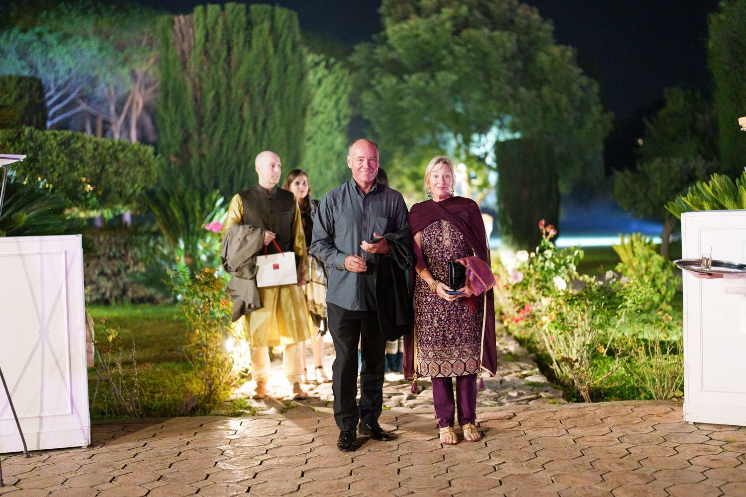Garden party scene with guests strolling along illuminated stone walkways.