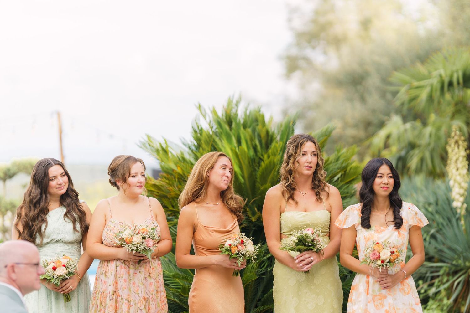 Bridesmaids in blush pink dresses hold bouquets while standing in a line at an outdoor wedding ceremony.