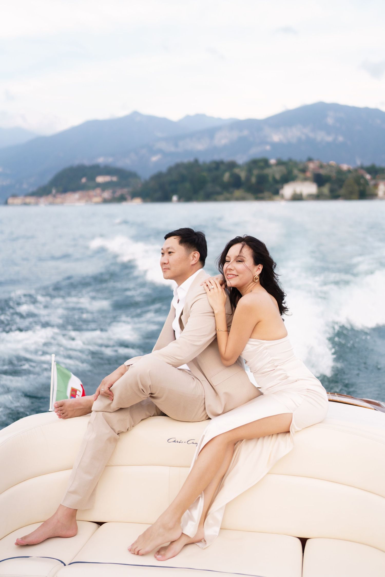 A couple relaxes together on the cushioned deck of a boat with mountains and ocean in the background.