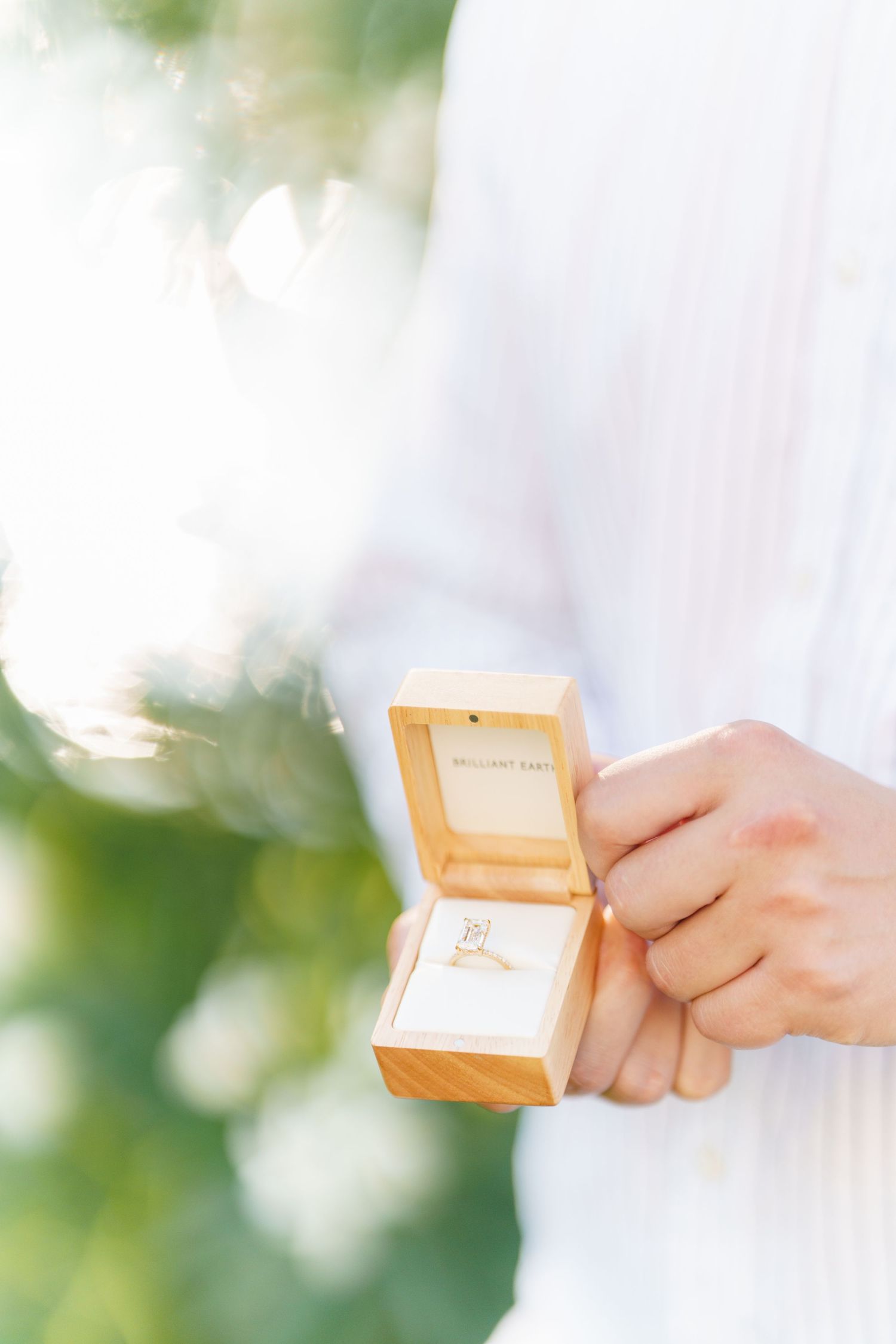 Hands holding a delicate peach-colored jewelry or ring box against a soft, dreamy bokeh background.