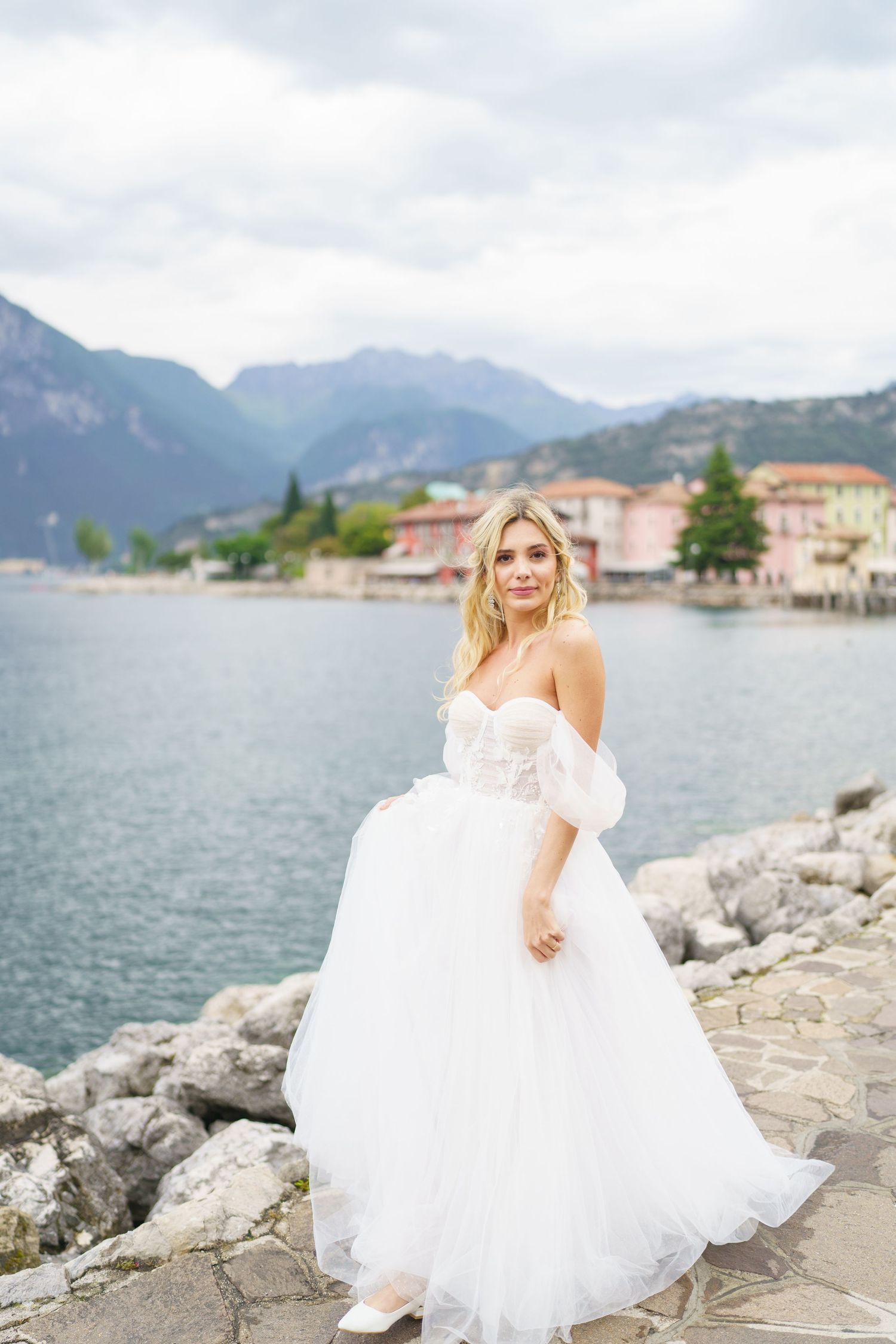 A bride in a flowing white wedding dress sits gracefully on rocky shores of Lake Como with mountains in the background.