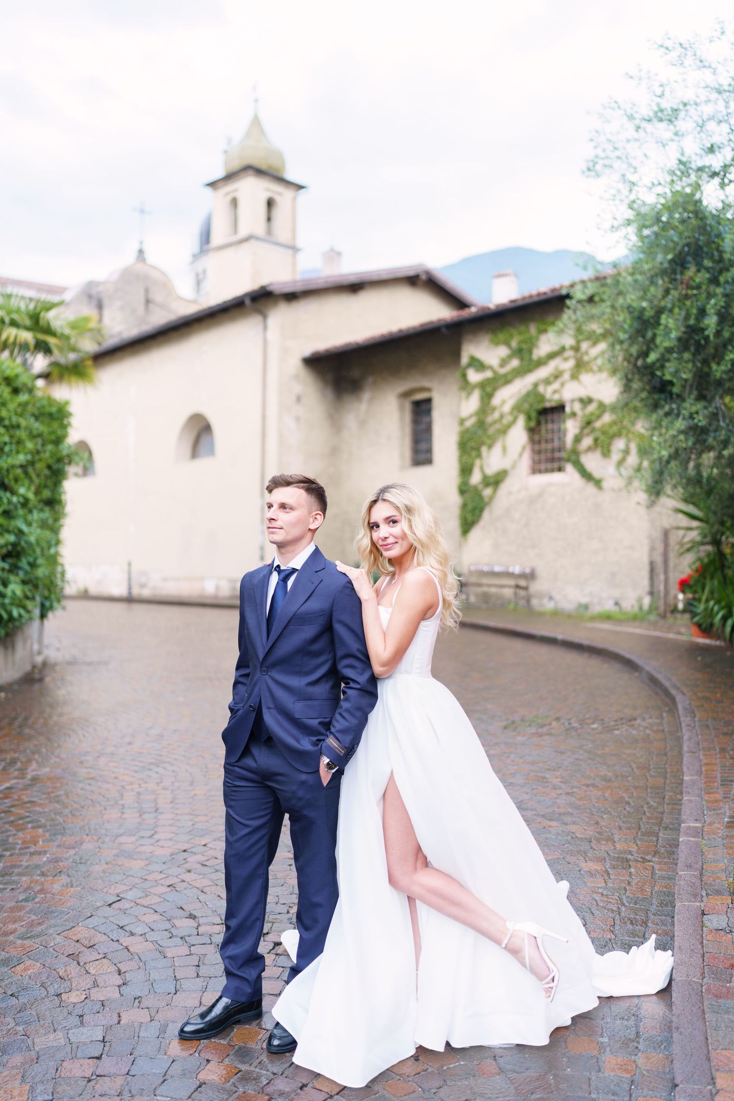 A couple in wedding attire poses playfully on cobblestone steps outside a historic church building.