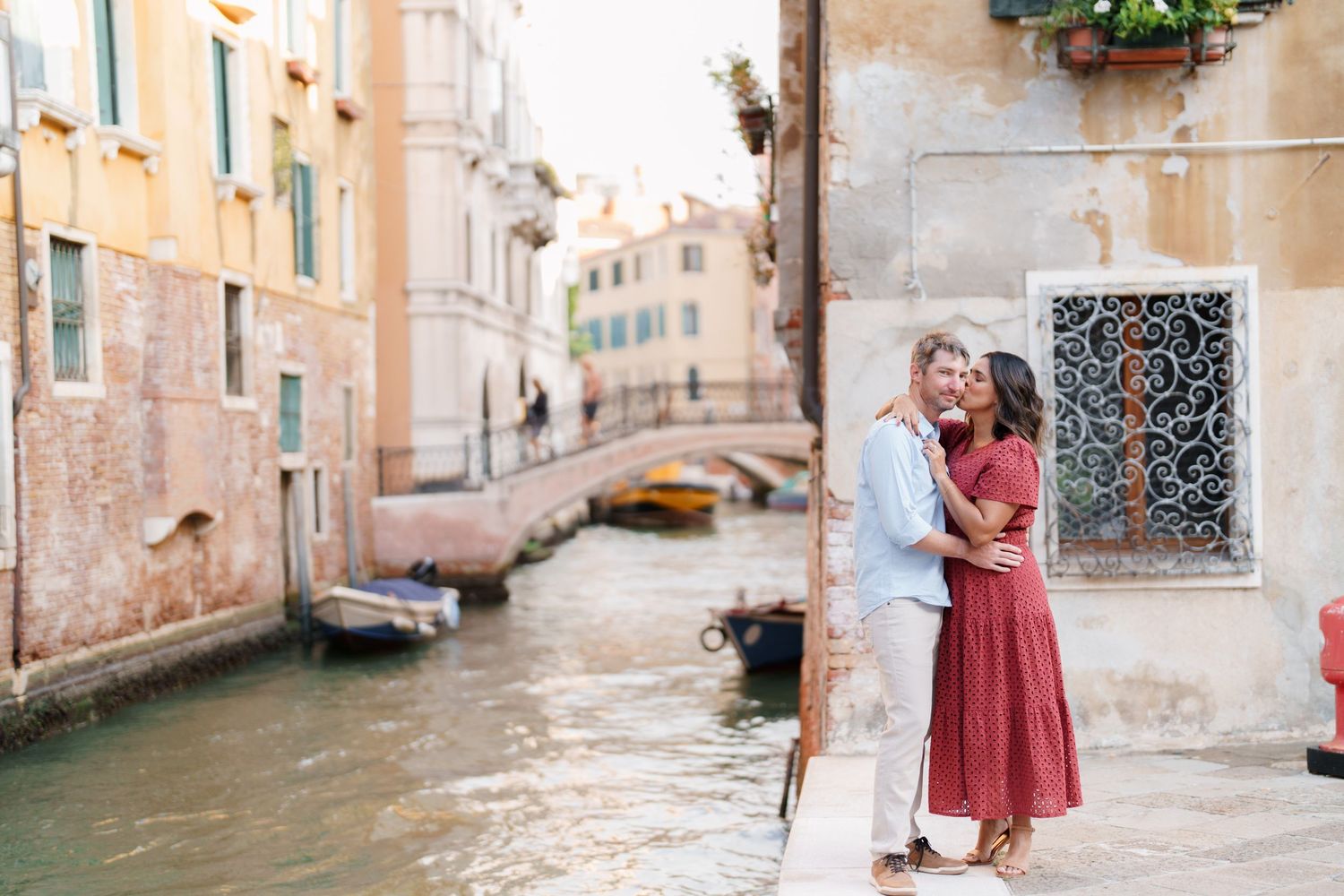 Romantic couple embracing on a Venice canal walkway, surrounded by colorful buildings and boats in Italy.