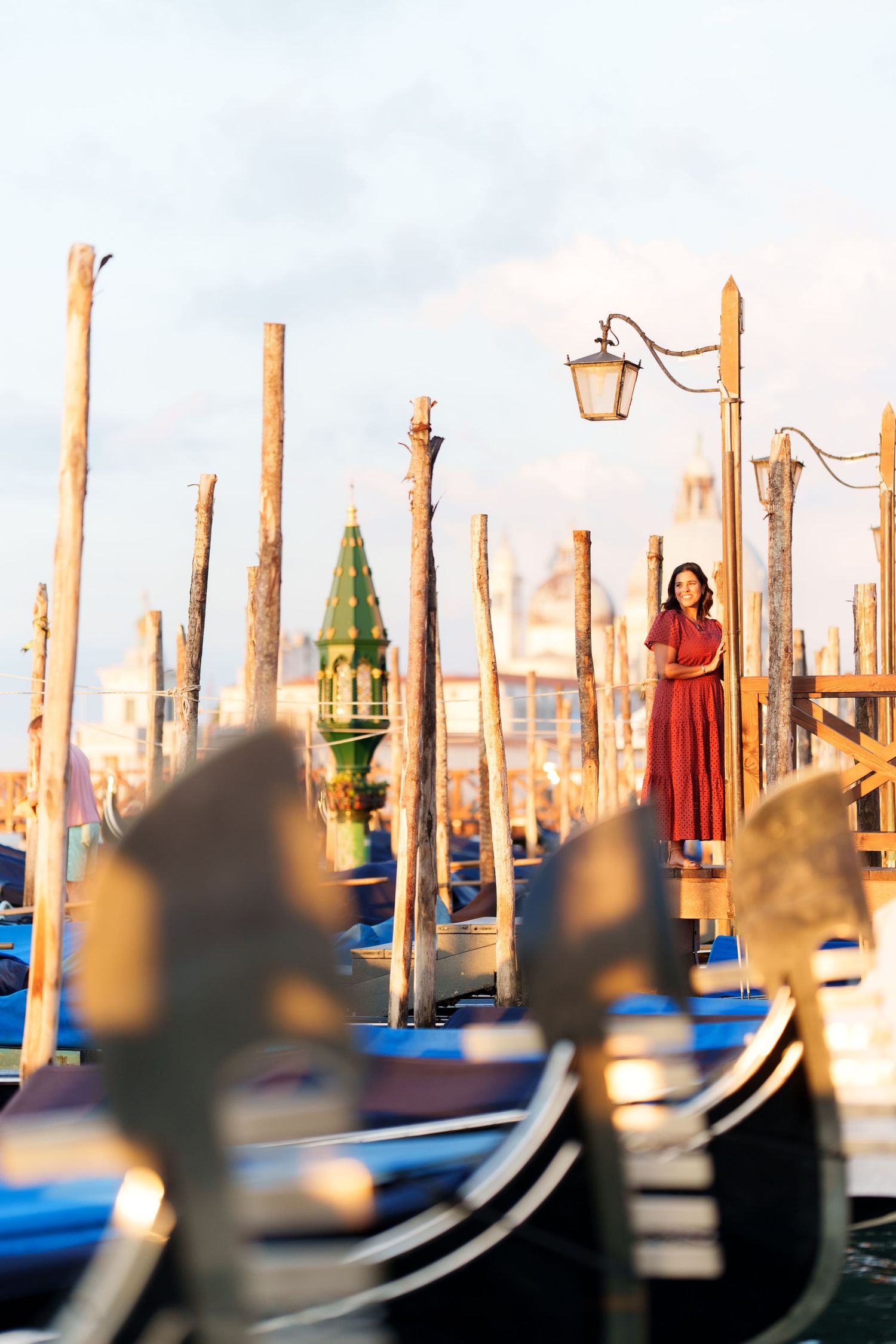 Woman in red dress standing among gondolas and wooden poles at Venice waterfront with soft golden light.