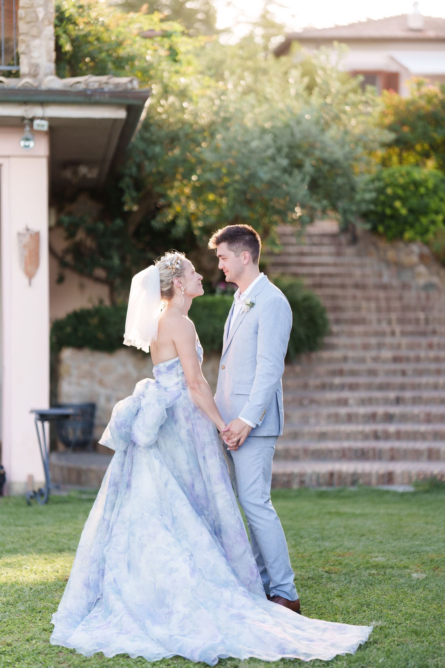 Bride in blue floral gown and groom in gray suit hold hands gazing at each other on green lawn at outdoor wedding.